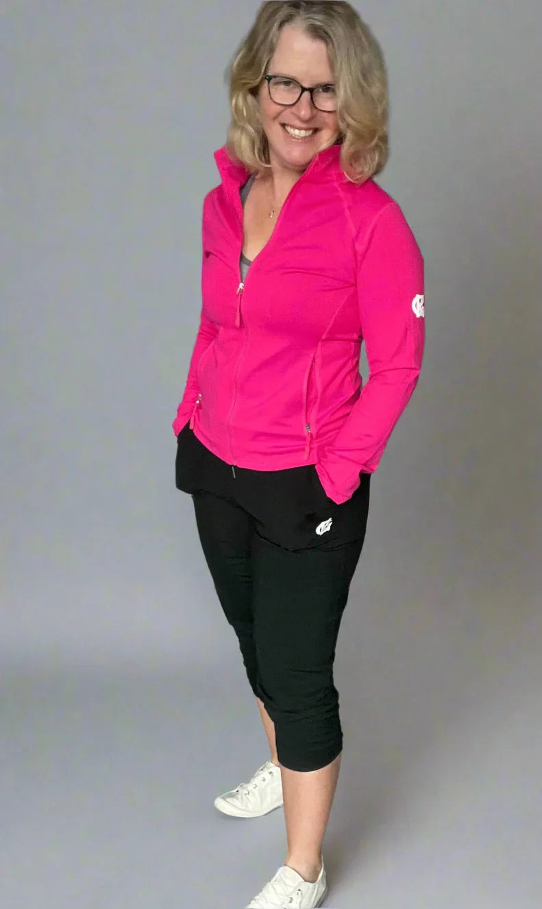 Woman in pink zip-up athletic jacket, black capri pants, white sneakers, smiling indoors
