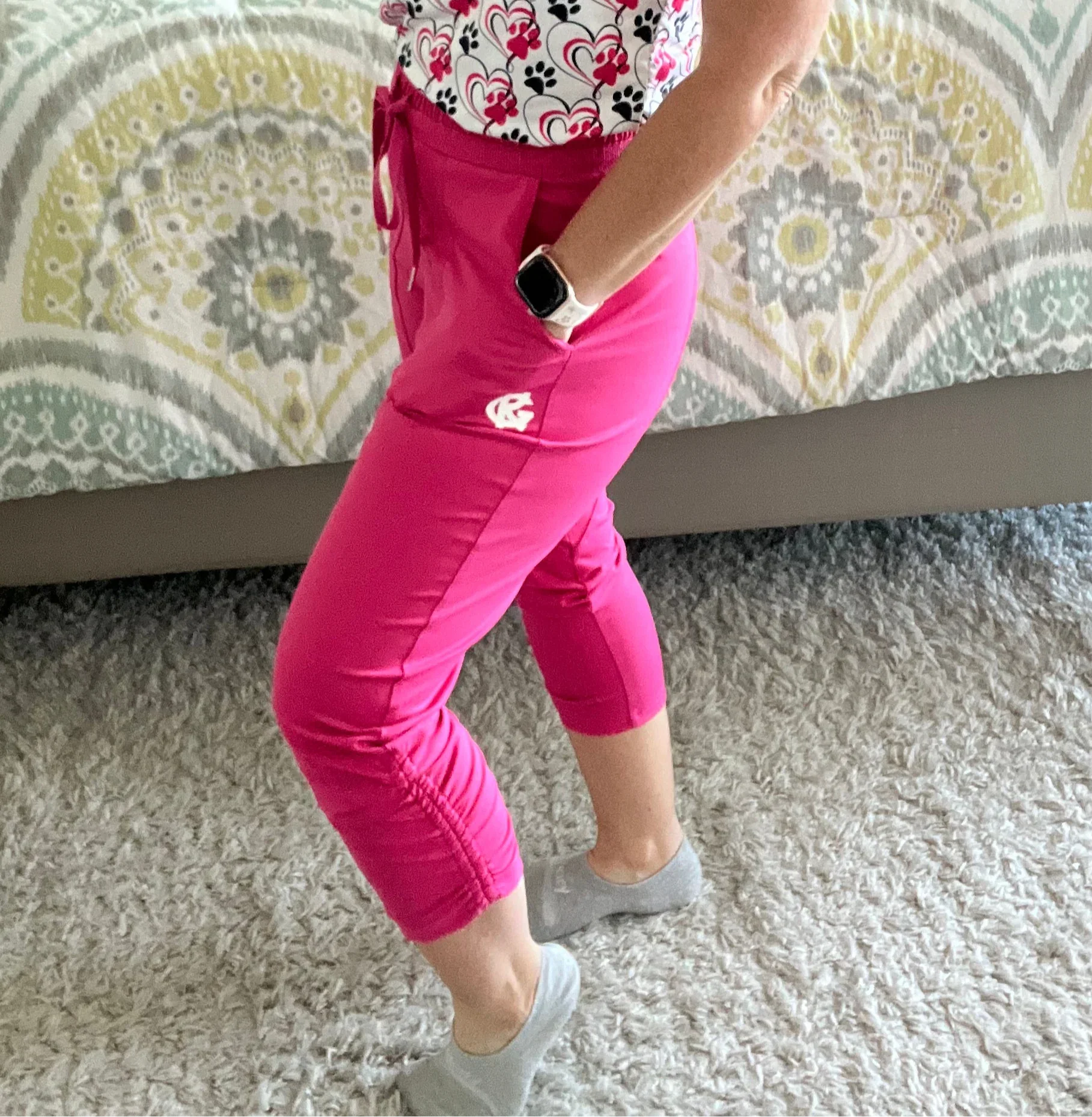 Woman wearing hot pink scrubs pants, heart and paw print top, smart watch, standing on carpet