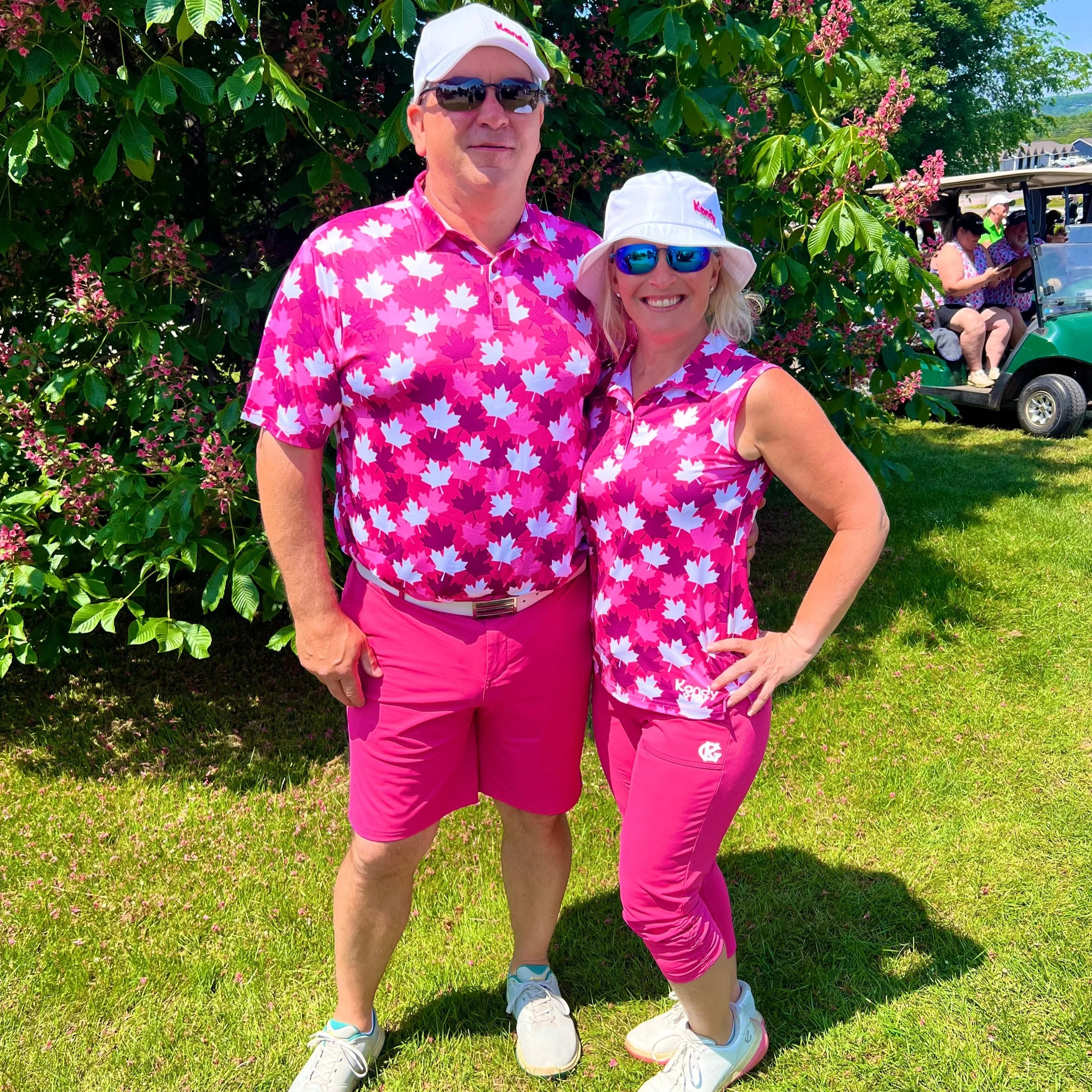 Two people in matching pink maple leaf golf outfits posing outdoors near a green golf cart.