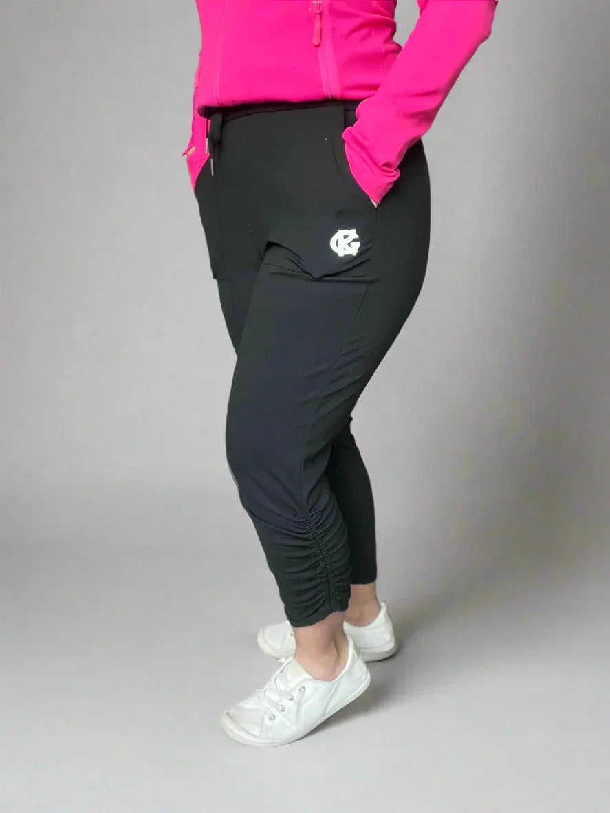 Woman in black jogger pants with side logo, pink jacket, and white sneakers standing indoors