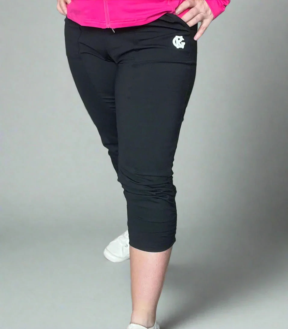 Woman wearing black capri leggings with logo, pink jacket, and white sneakers on gray background