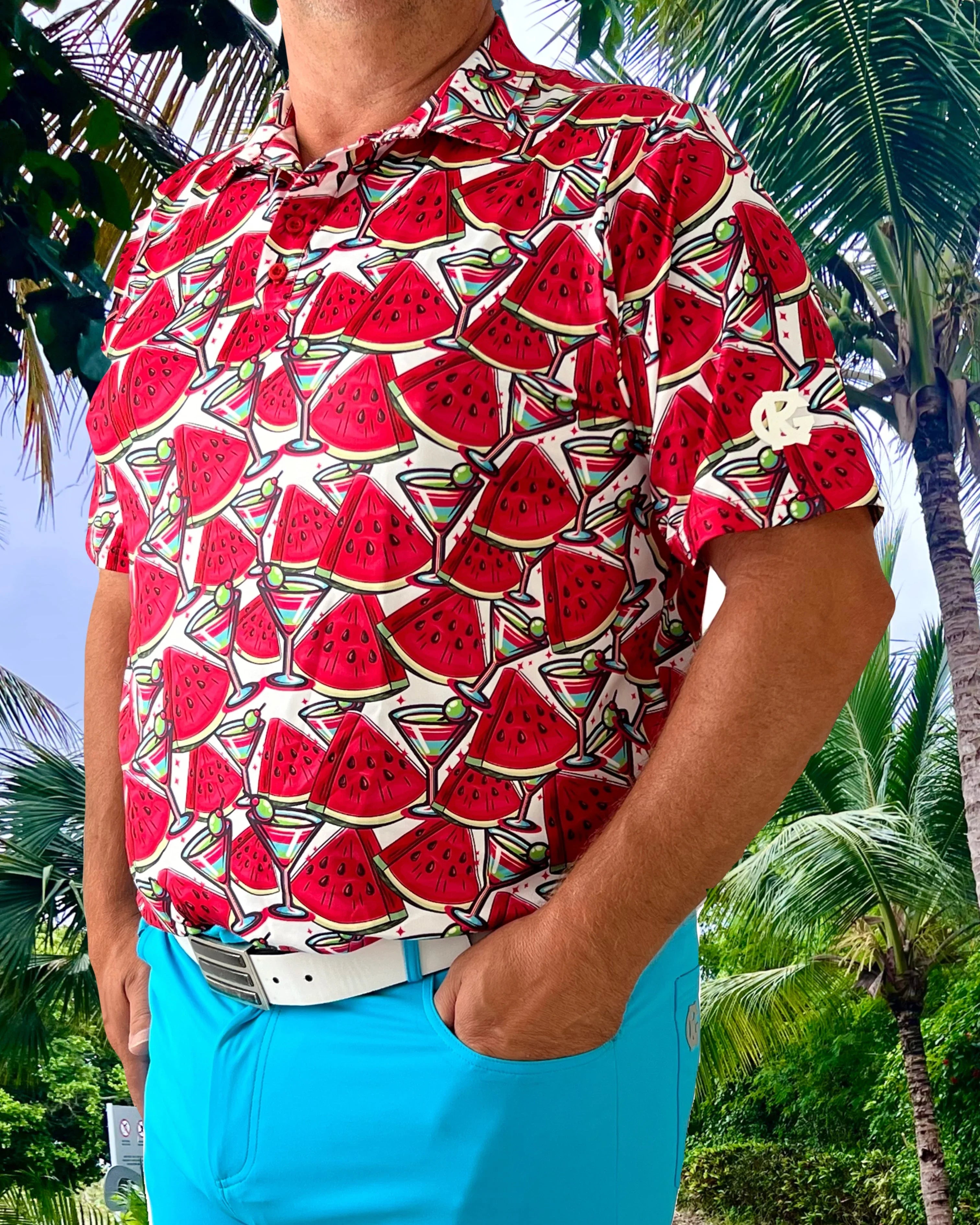 Man in watermelon print shirt and blue shorts standing outdoors with palm trees