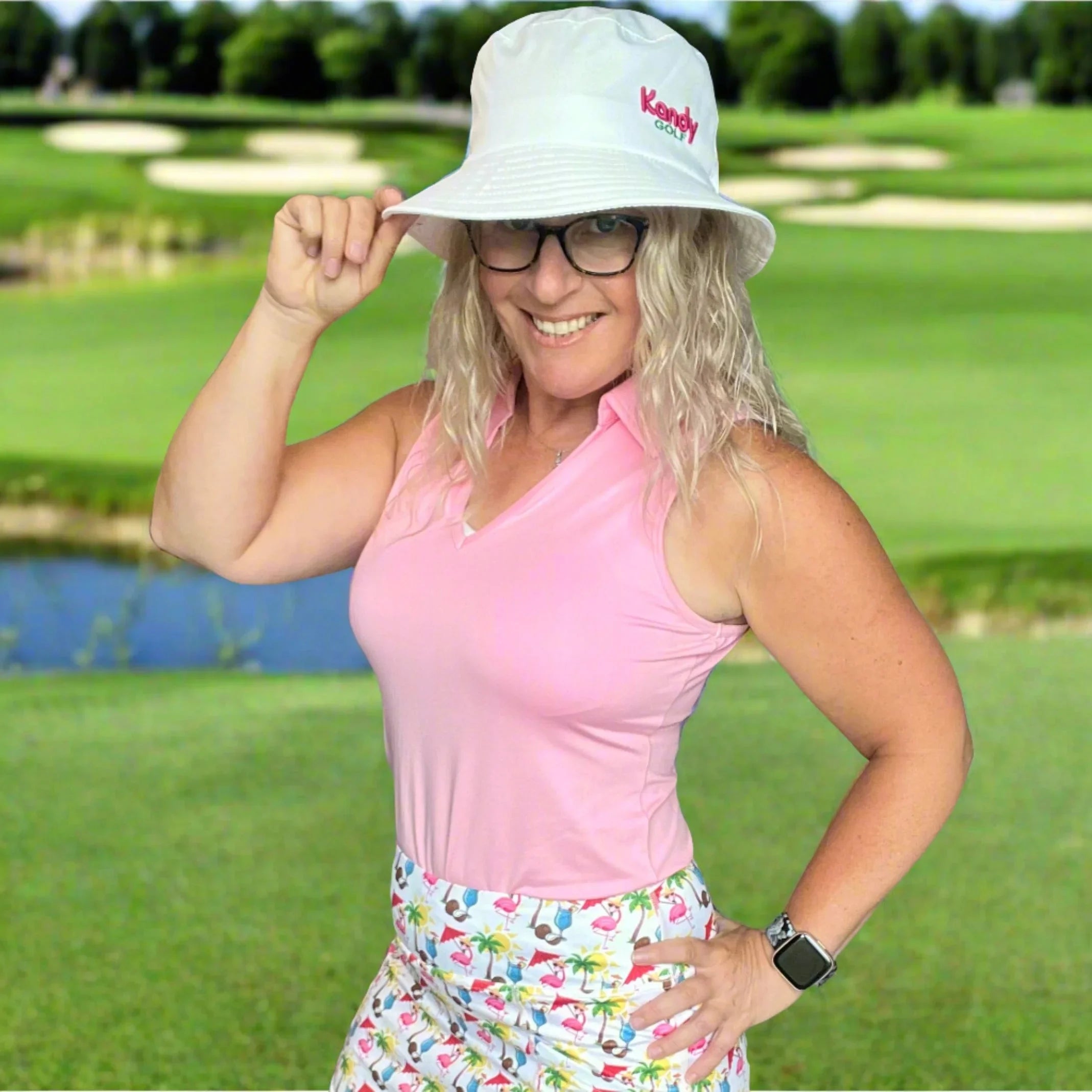 Smiling woman in pink golf top, printed skirt, and Kendly Golf bucket hat on a golf course