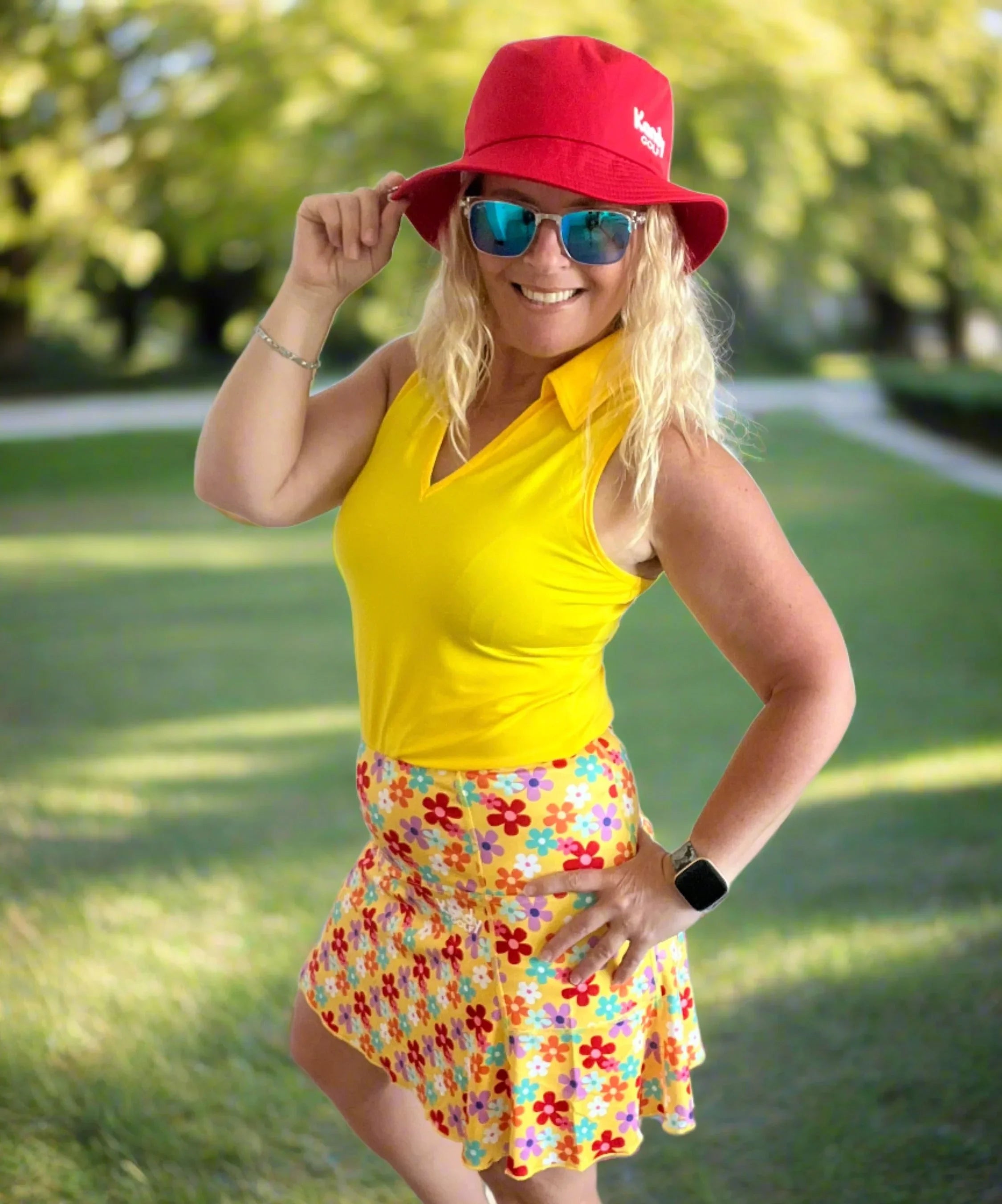 Woman in yellow sleeveless golf top, colorful floral skirt, red hat, and sunglasses outdoors