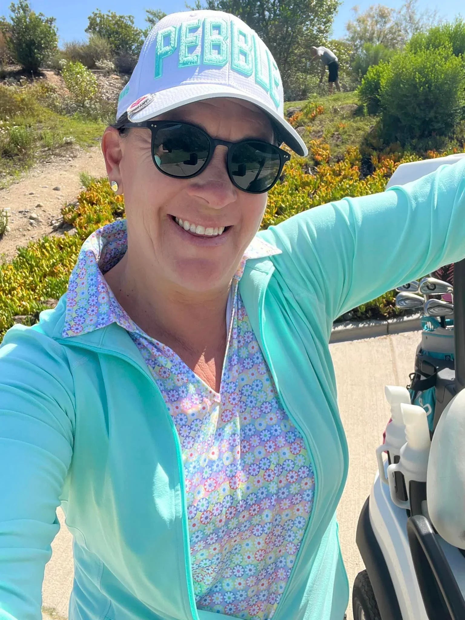 Woman in sunglasses and PEBBLE hat smiling on sunny golf course beside golf cart