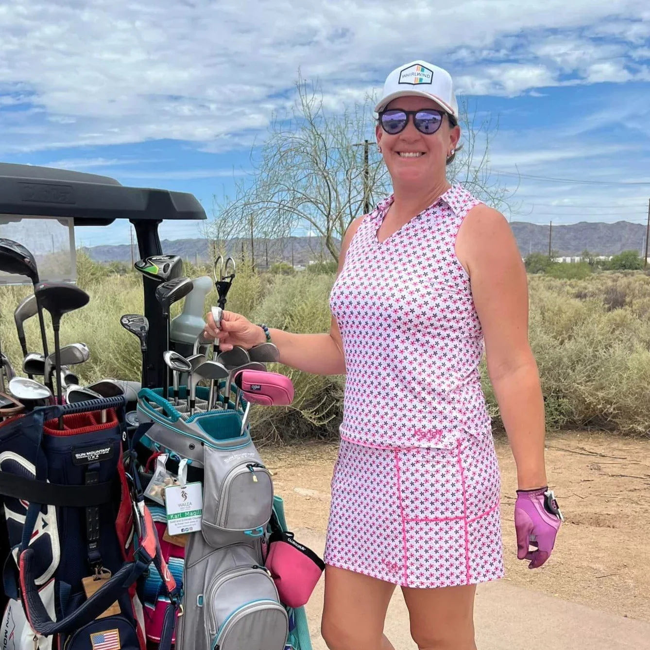 Smiling woman golfer in pink polka dot outfit standing by golf cart with clubs outdoors