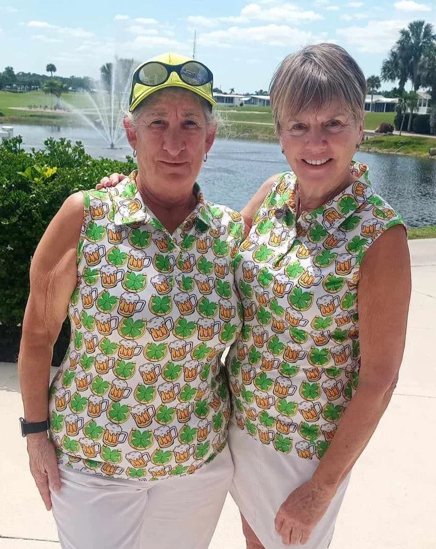 Two women in matching shamrock and beer print shirts by a pond with fountain, palm trees, sunny day