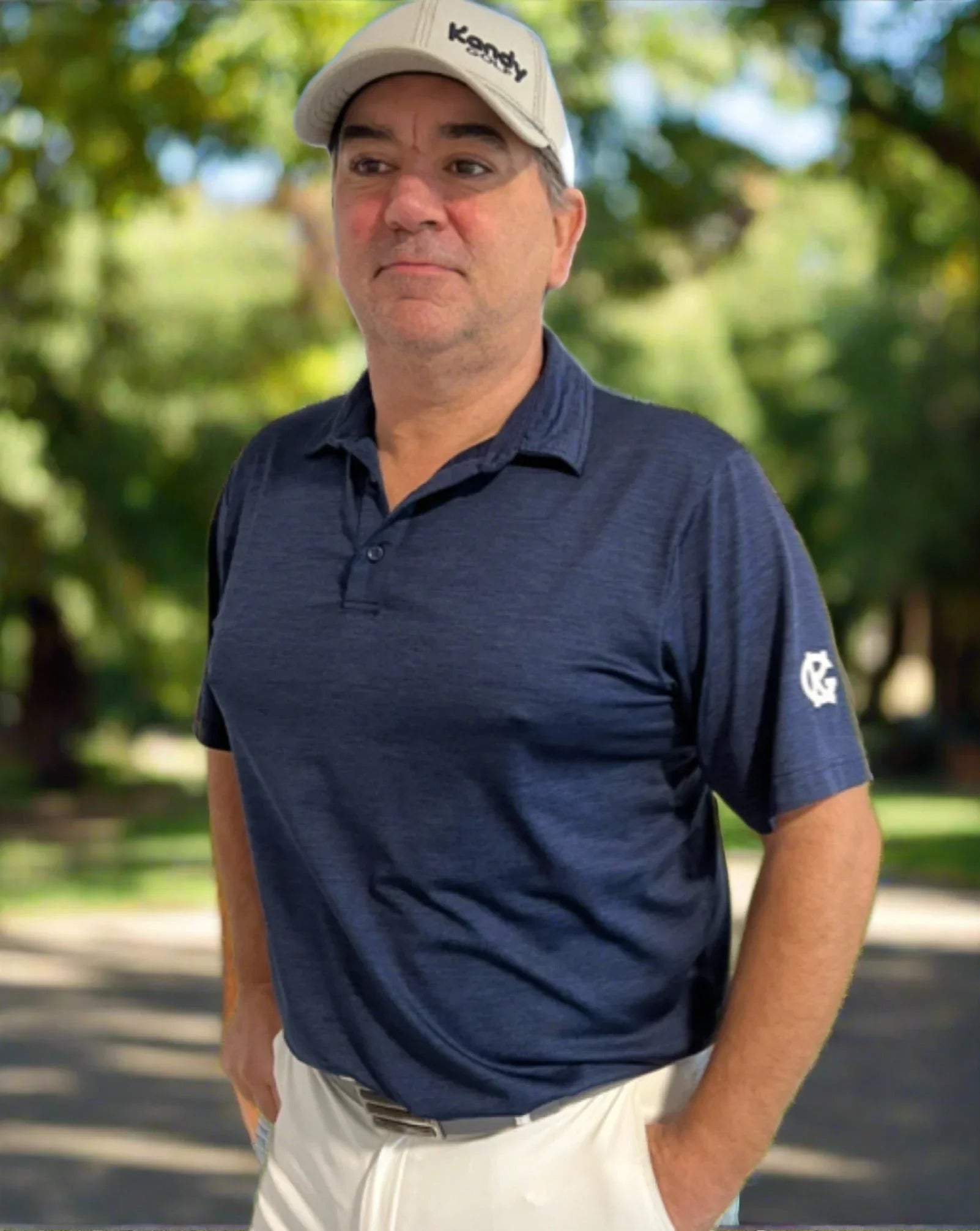 Man in navy golf polo and tan cap standing outdoors in a sunny park setting