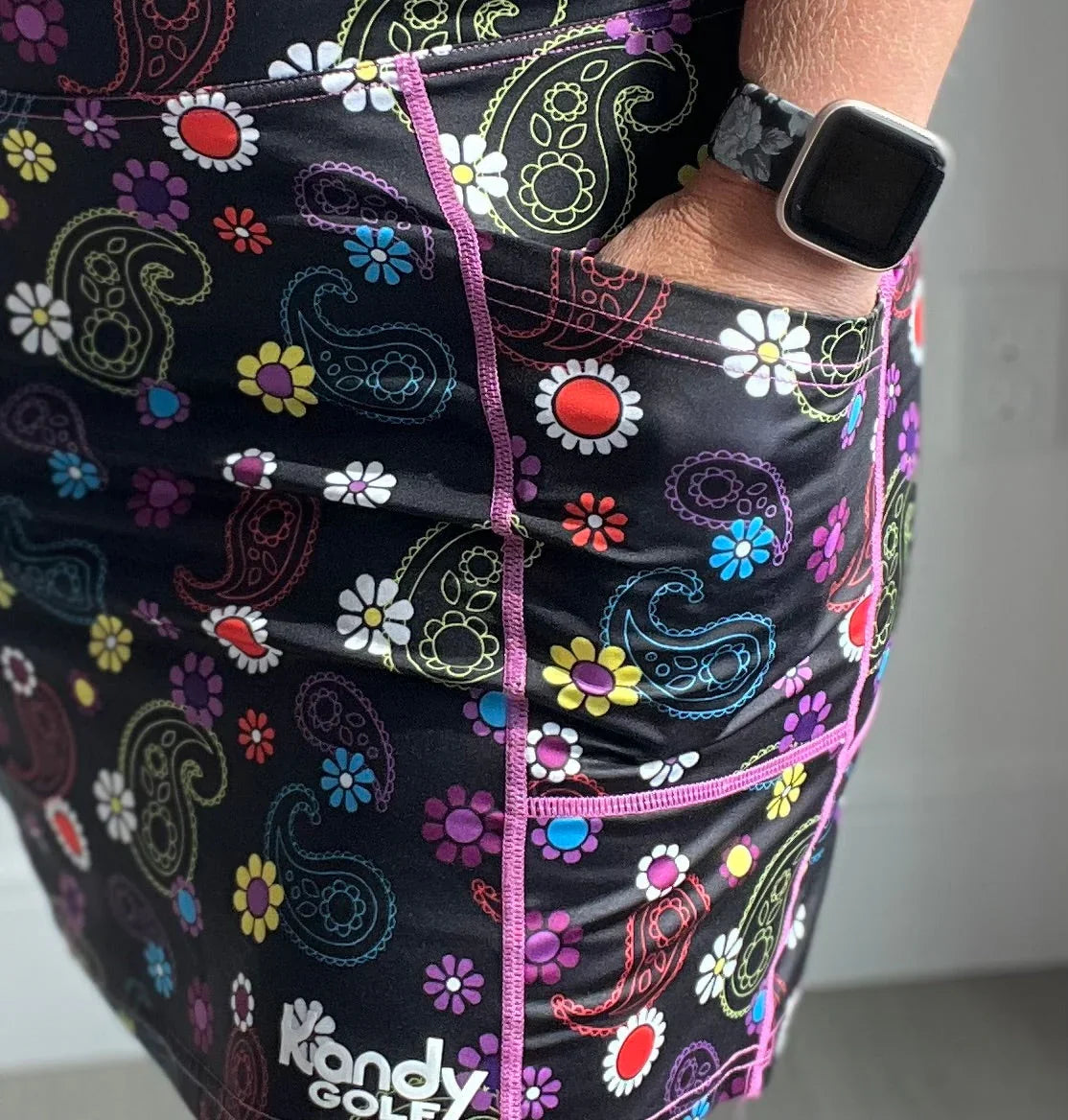 Colorful paisley and daisy print golf skirt with a pocket, Kandy Golf logo, smartwatch.