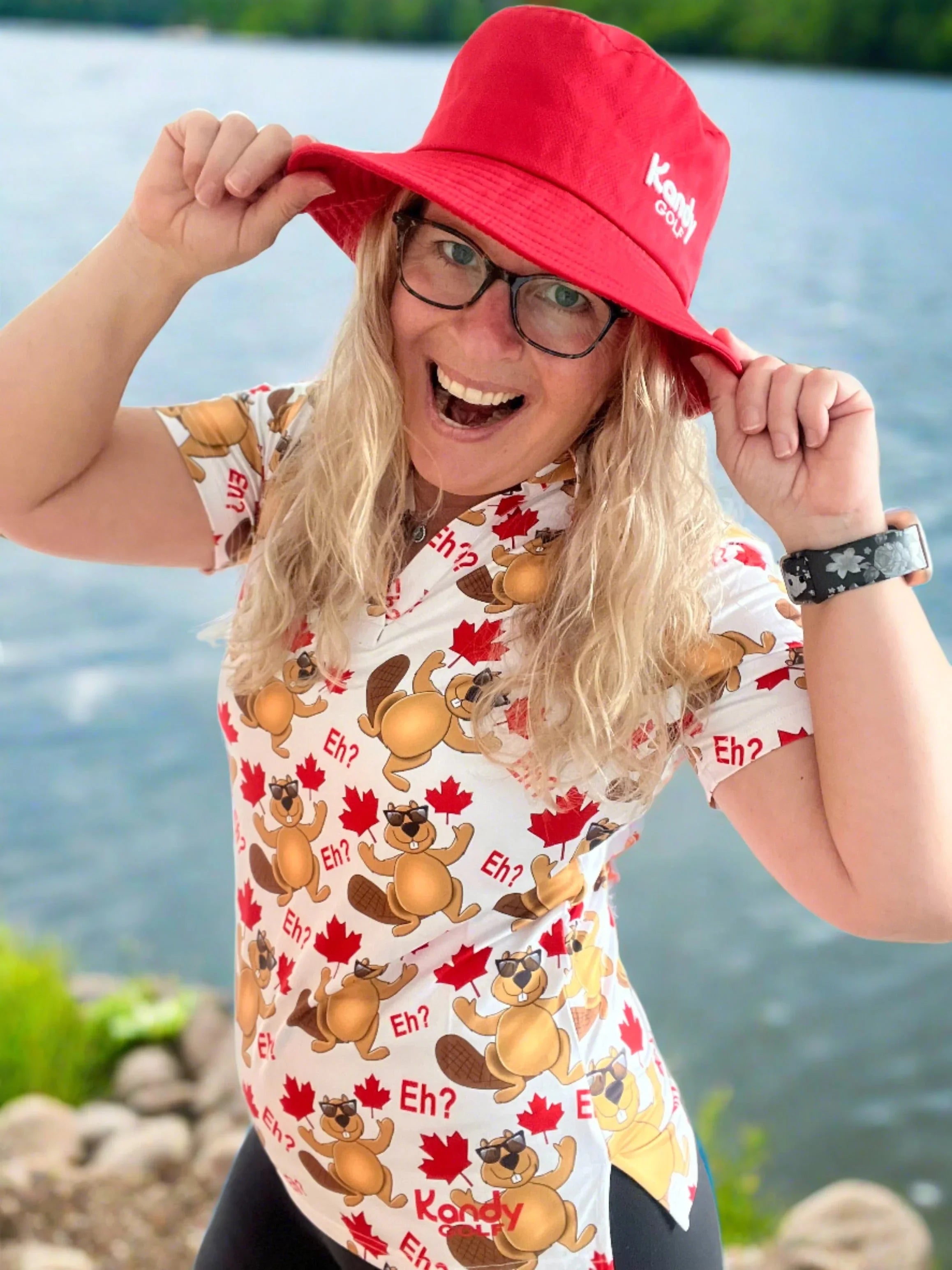 Smiling woman in red Kandy Golf hat and Canada-themed shirt with beavers by a lake