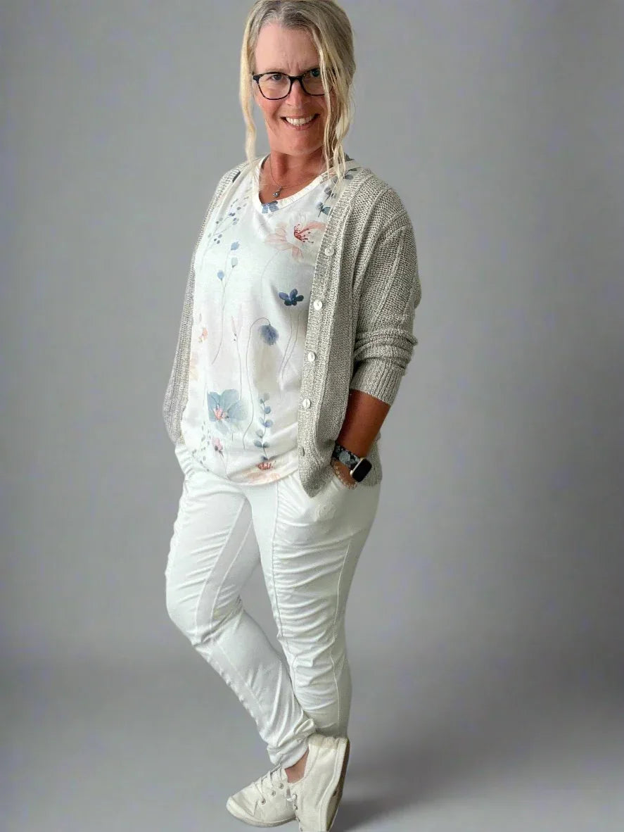 Smiling woman in glasses, light cardigan, floral shirt, white pants, and white sneakers