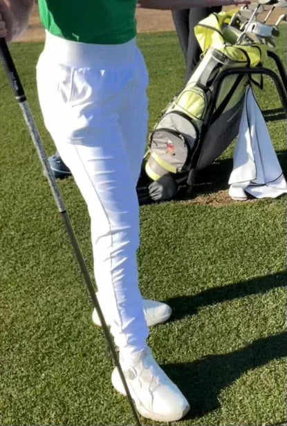 Golfer in white pants and green shirt on golf course with golf bag and clubs in background