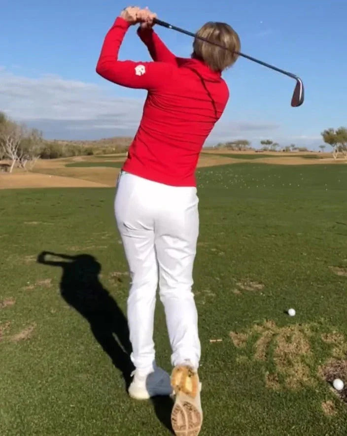 Golfer in red jacket and white pants swings club on sunny golf course fairway