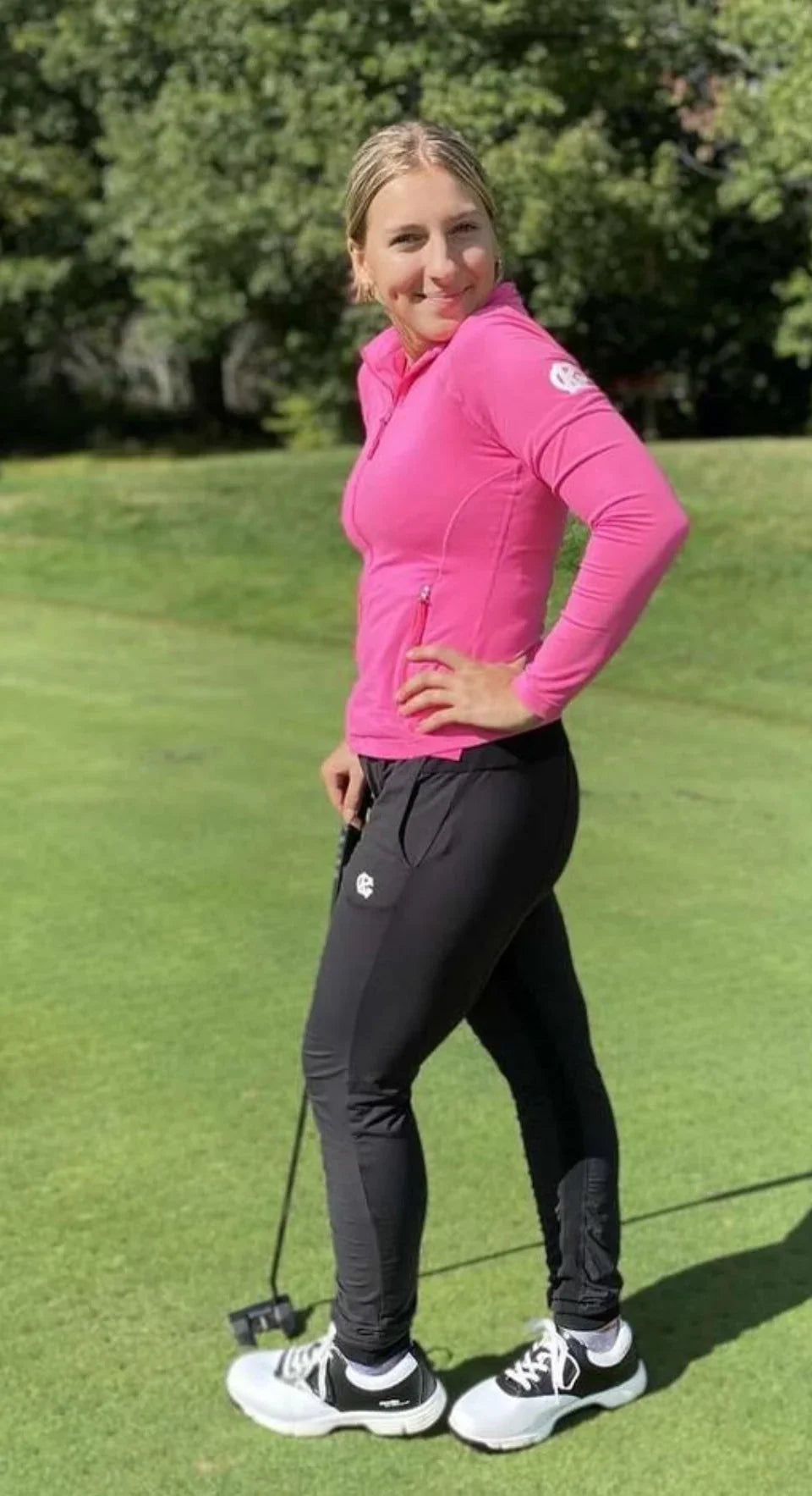 Woman golfer in pink jacket and black pants holding putter on green golf course
