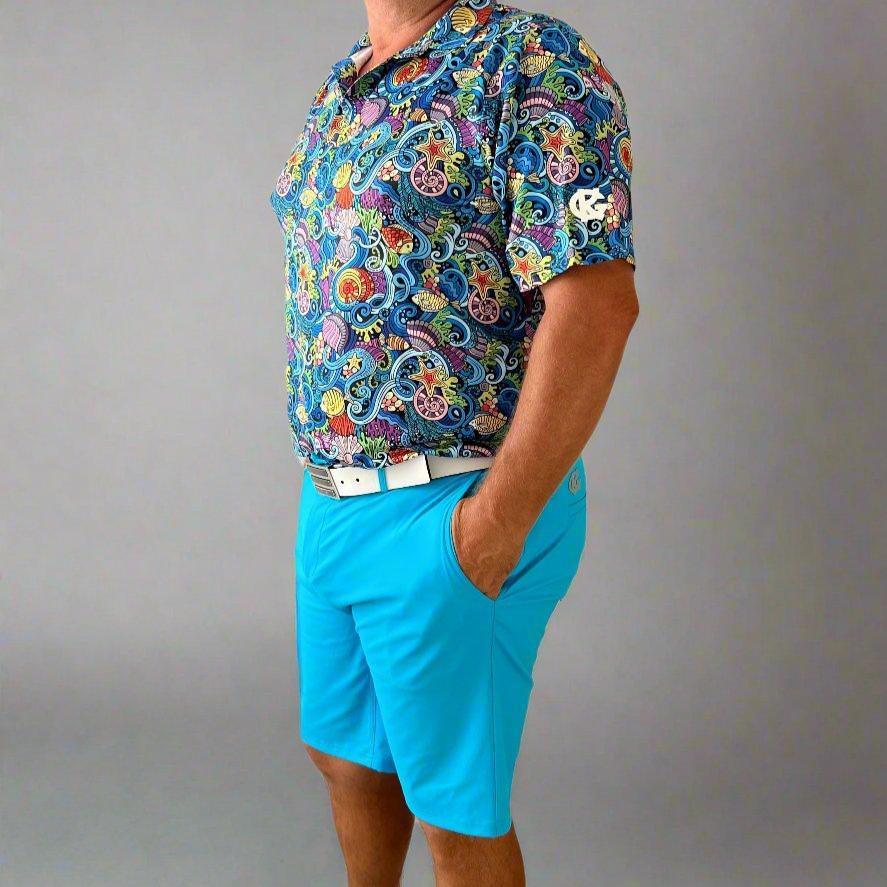Man wearing a colorful paisley shirt and bright blue shorts with a white belt, studio background