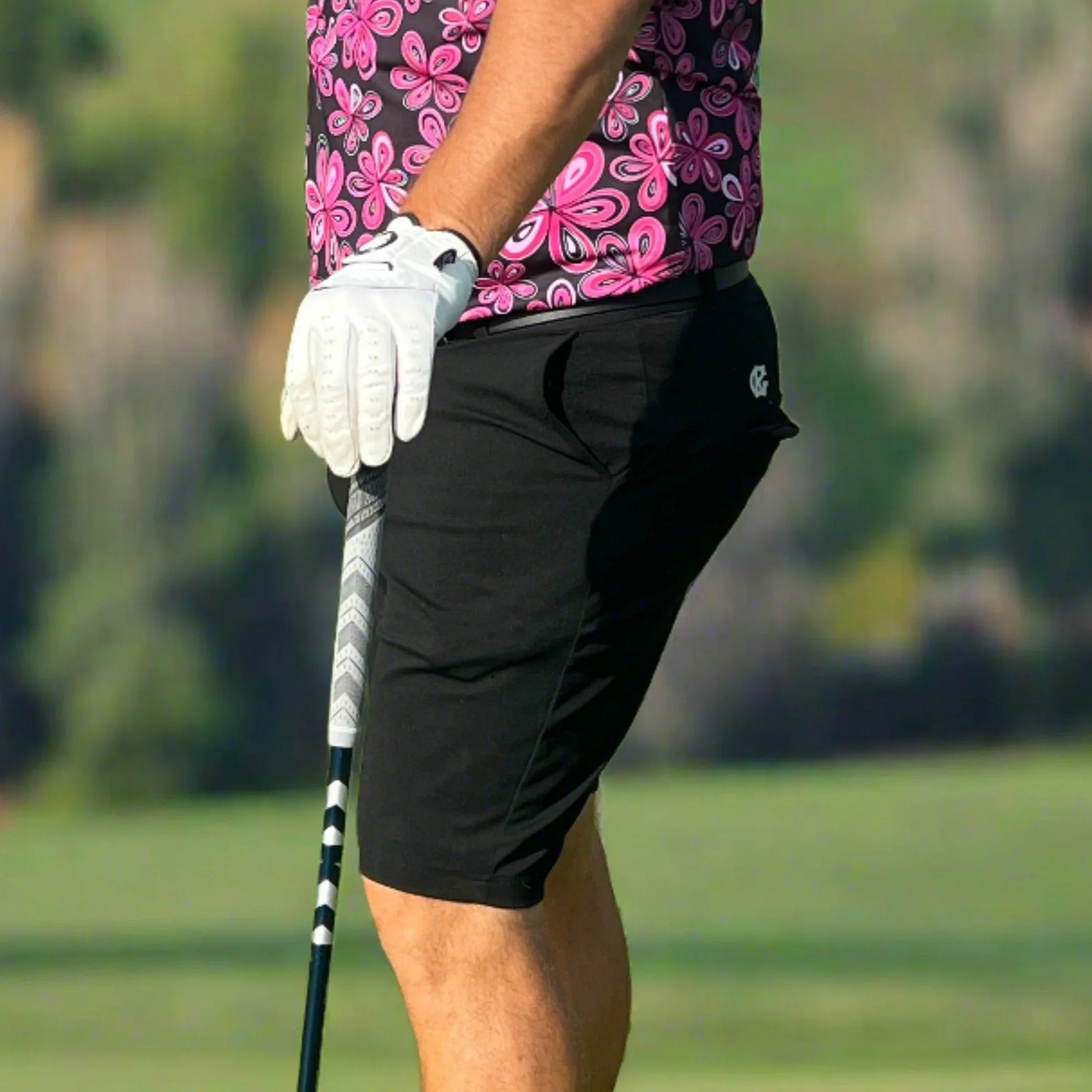 Golfer wearing black shorts, pink floral shirt, and white glove holding a golf club outdoors