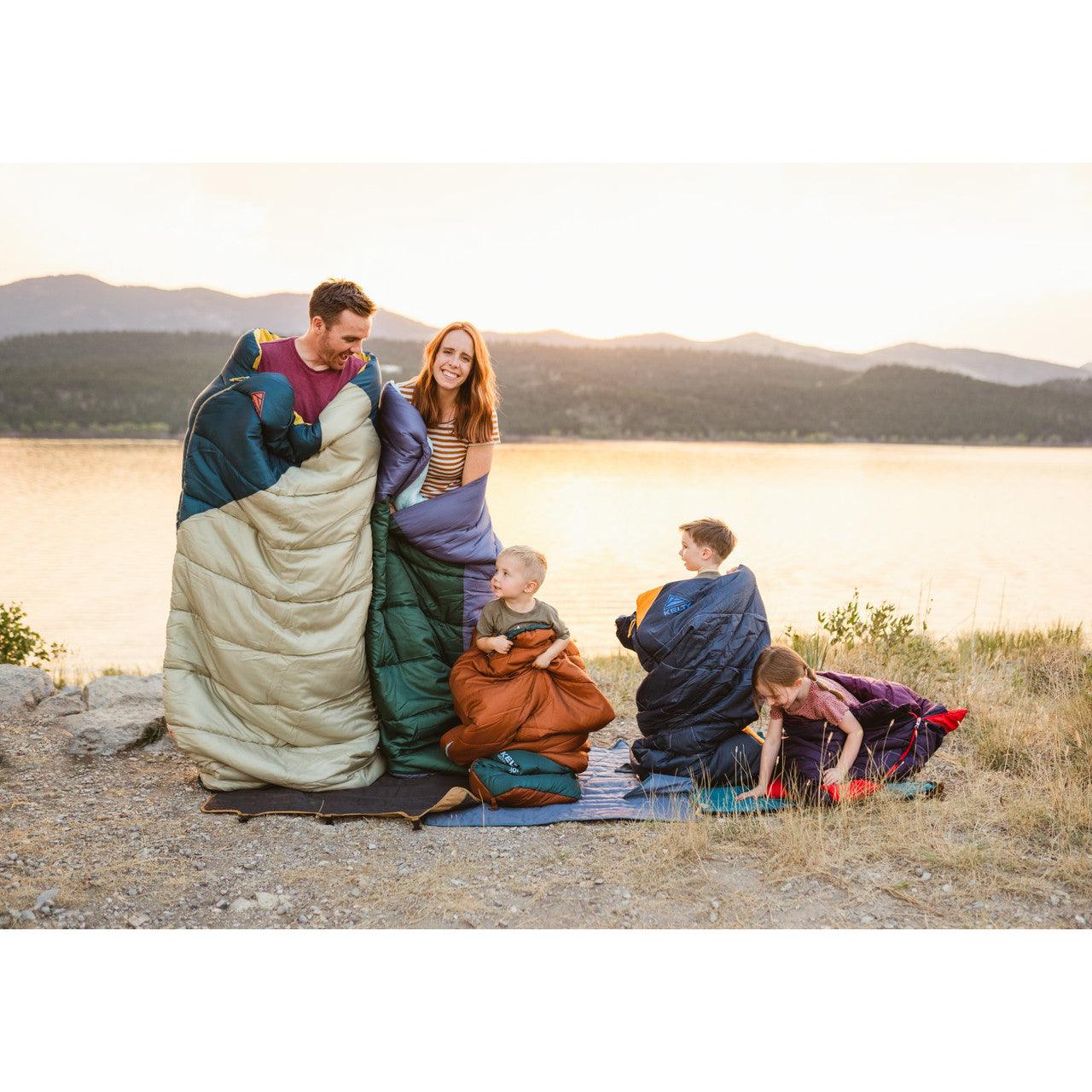 Family in sleeping bags camping by a lake at sunset with mountains in background
