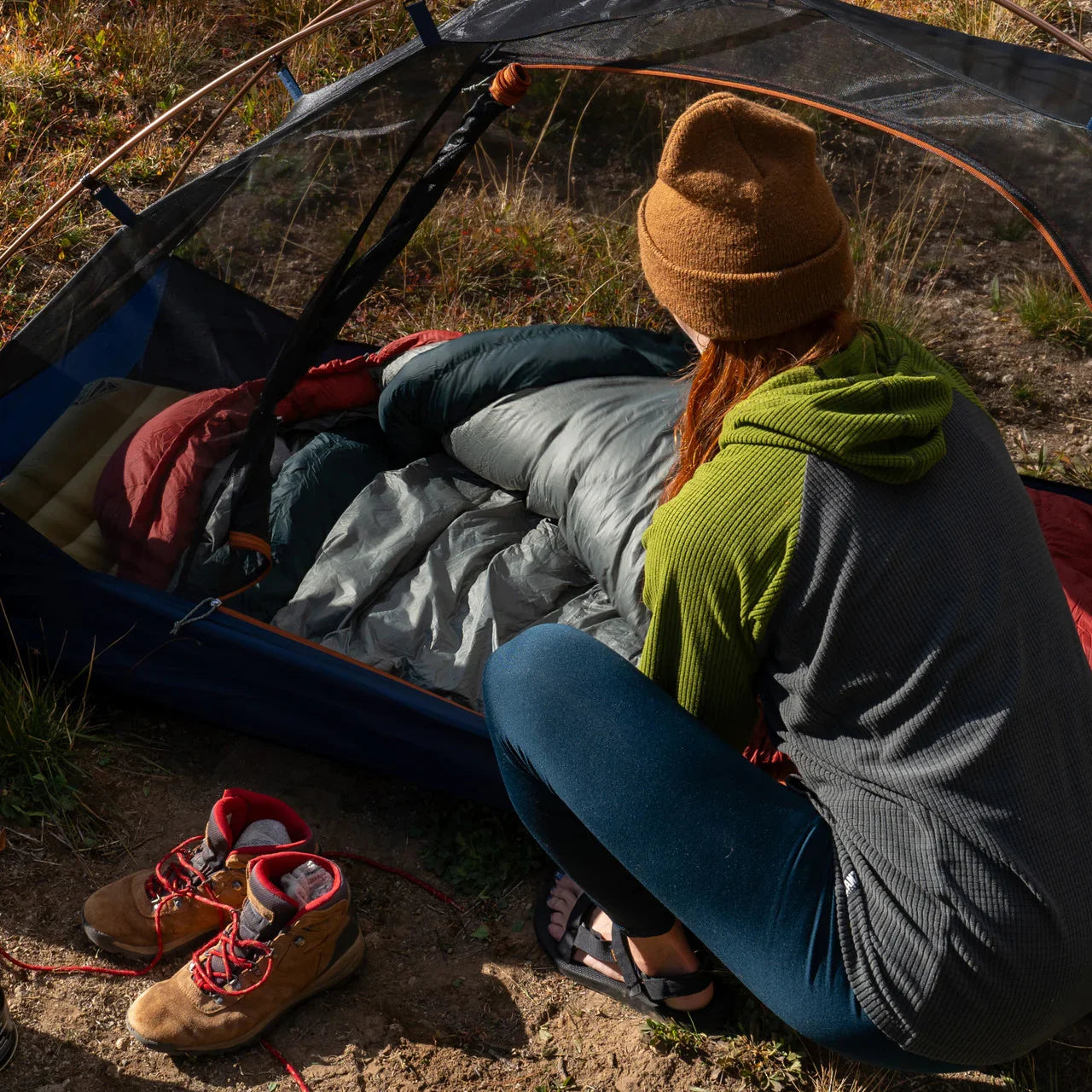 Camper in beanie and hoodie arranging sleeping bag inside tent, hiking boots outside on grass