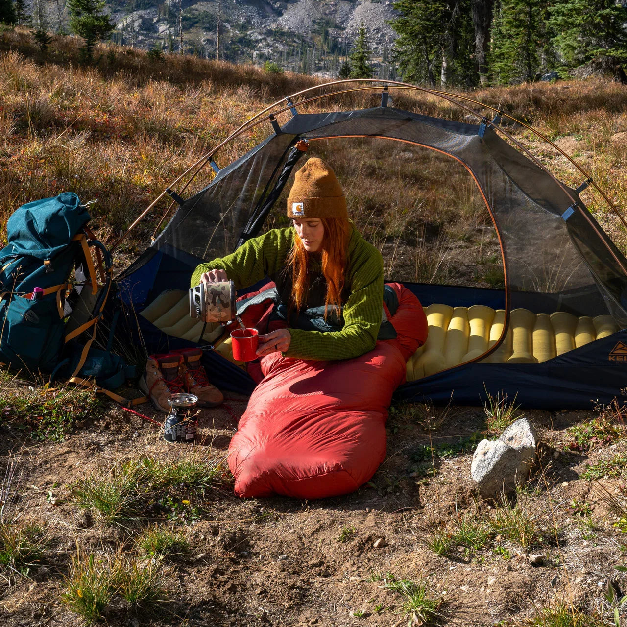 Woman camping in a tent, pouring coffee, in a sleeping bag on a mountain meadow