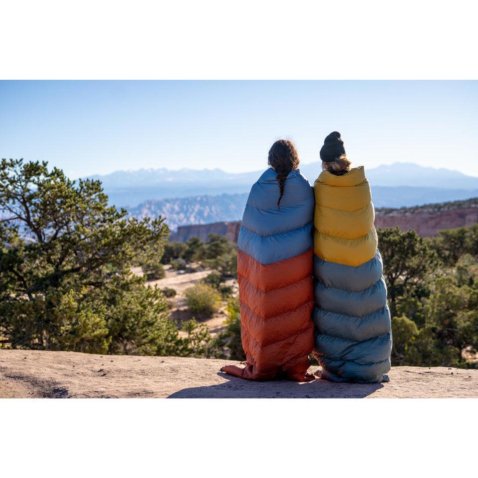 Two people in colorful sleeping bags overlook a scenic canyon landscape with distant mountains.