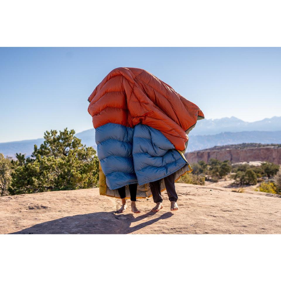 Two people covered by a red and blue puffy sleeping bag outdoors on a rocky overlook