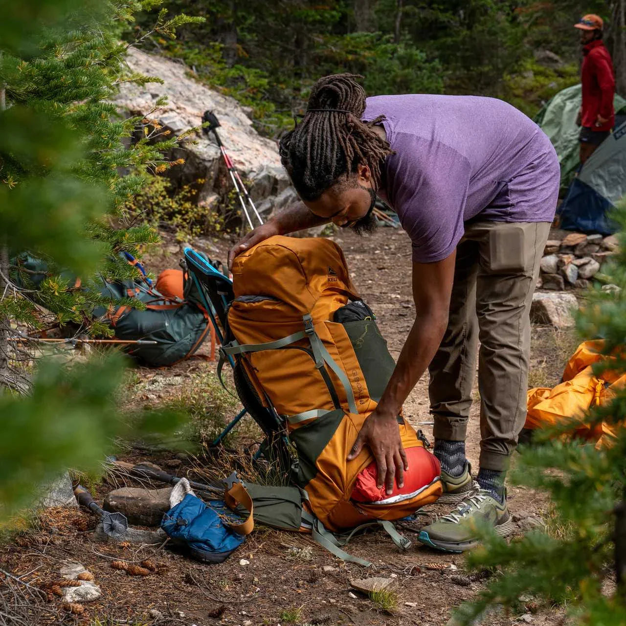 Man packing orange Kelty Glendale 65L backpack at forest campsite with tents and gear