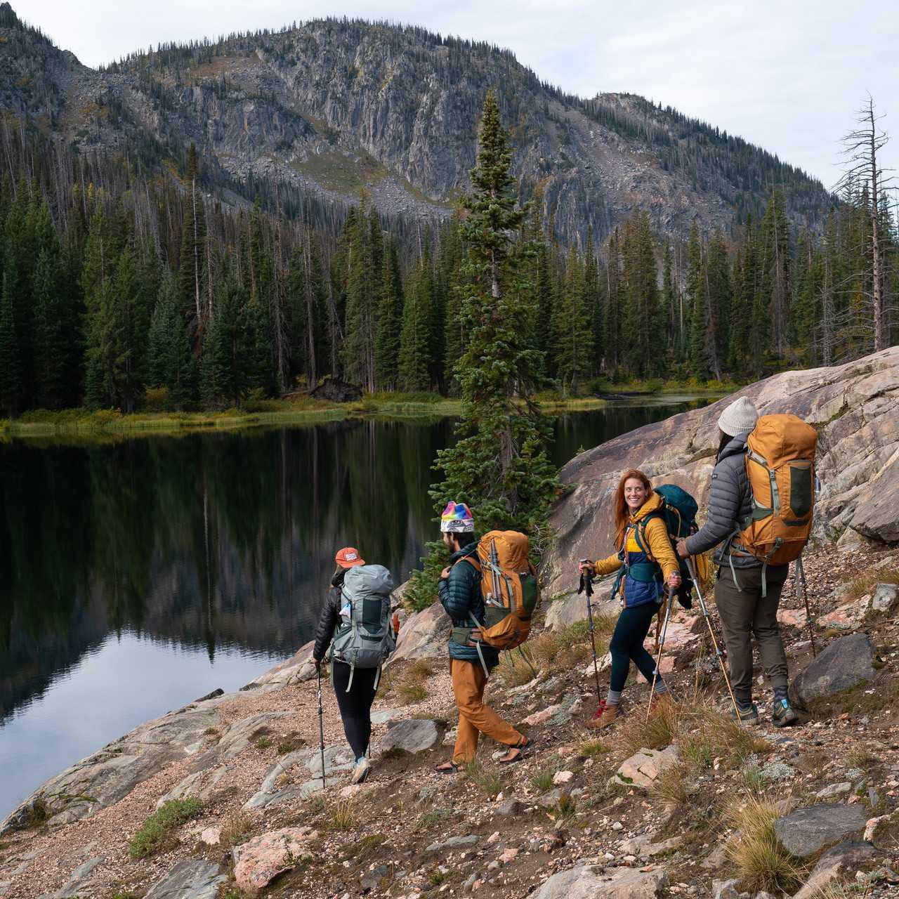 Hikers with backpacks walking on rocky lakeside trail in the mountains, forest in background.