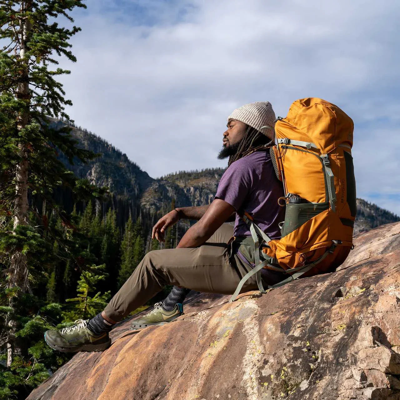 Hiker wearing yellow Kelty Glendale 65L backpack sitting on rocky mountain with trees.