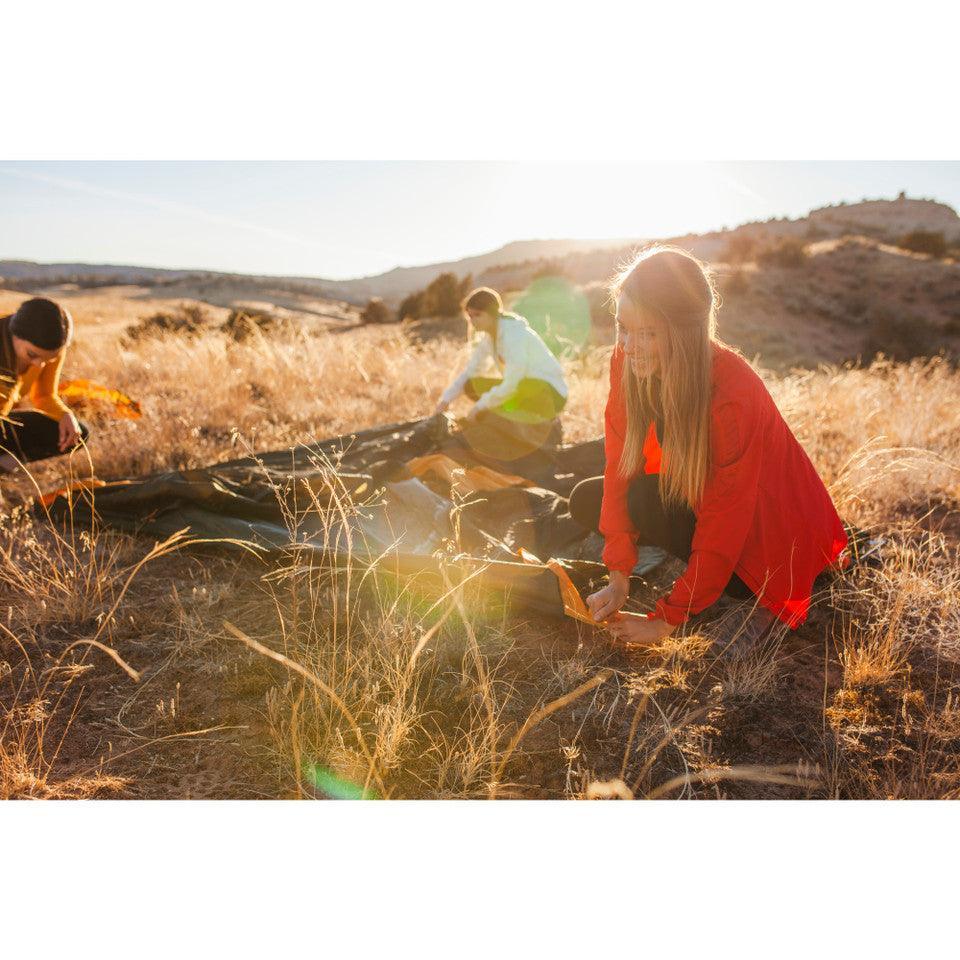 Three people setting up a tent in a sunny, dry grassy field with hills in the background.