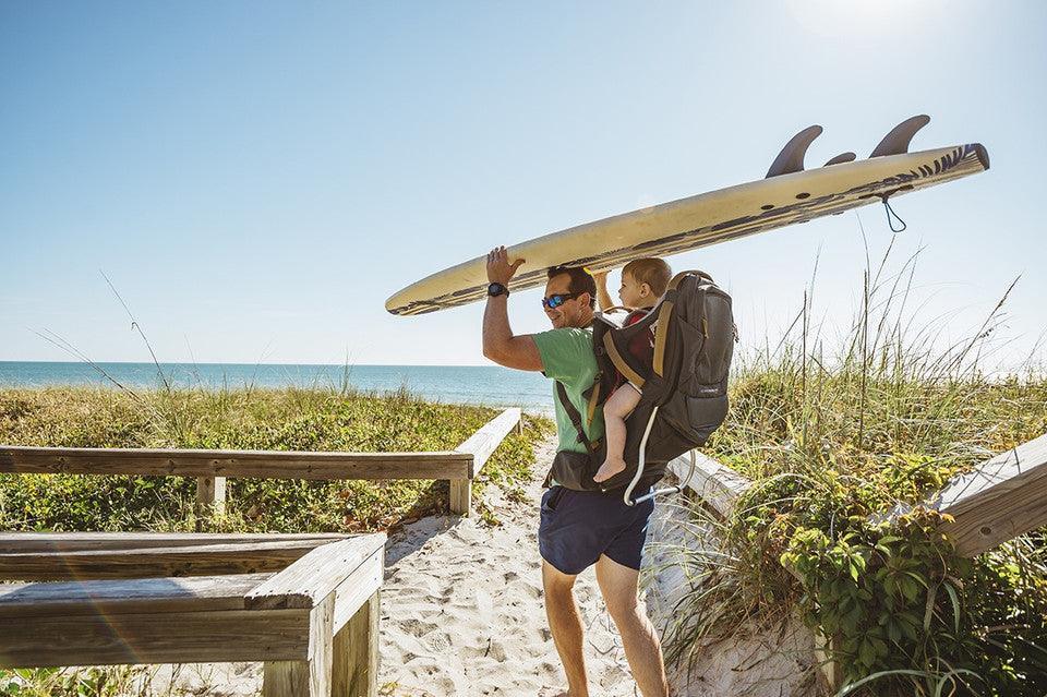 Man carrying surfboard and baby in backpack on sandy Florida beach path