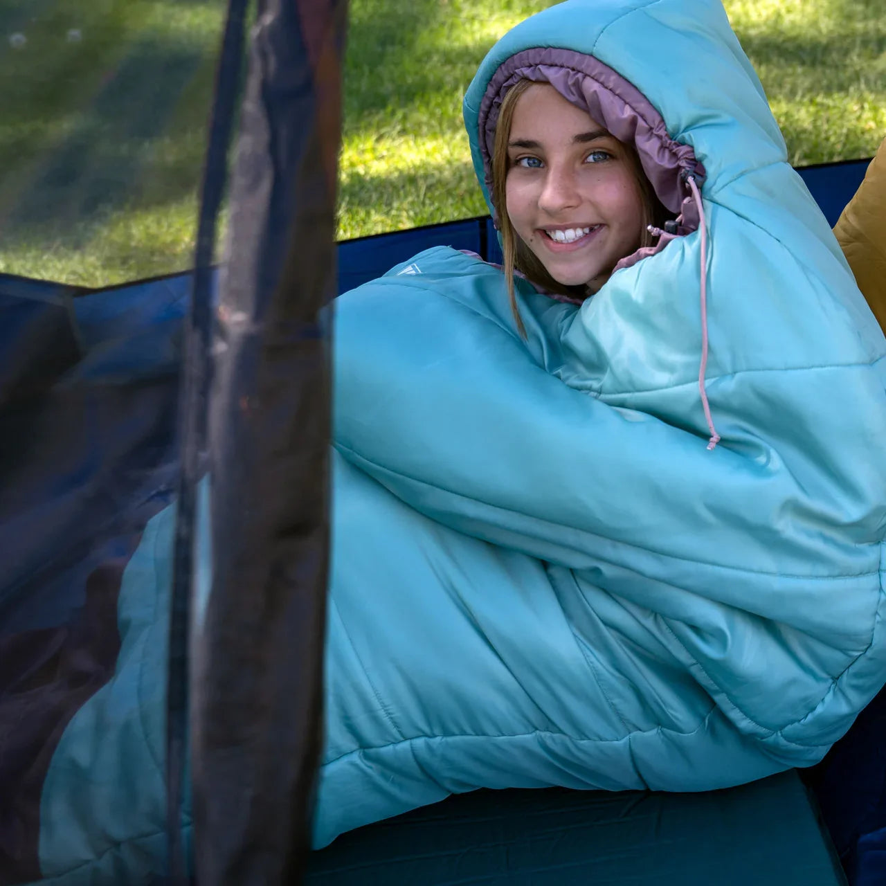 Smiling girl in blue hooded sleeping bag sitting inside a tent on grass