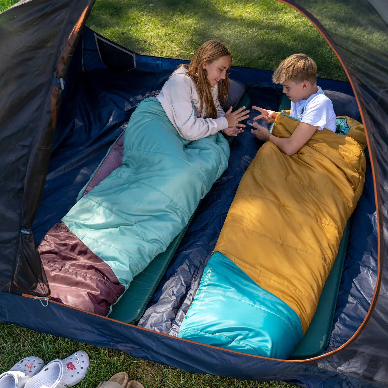 Two kids in colorful sleeping bags inside a tent, camping outdoors on grass