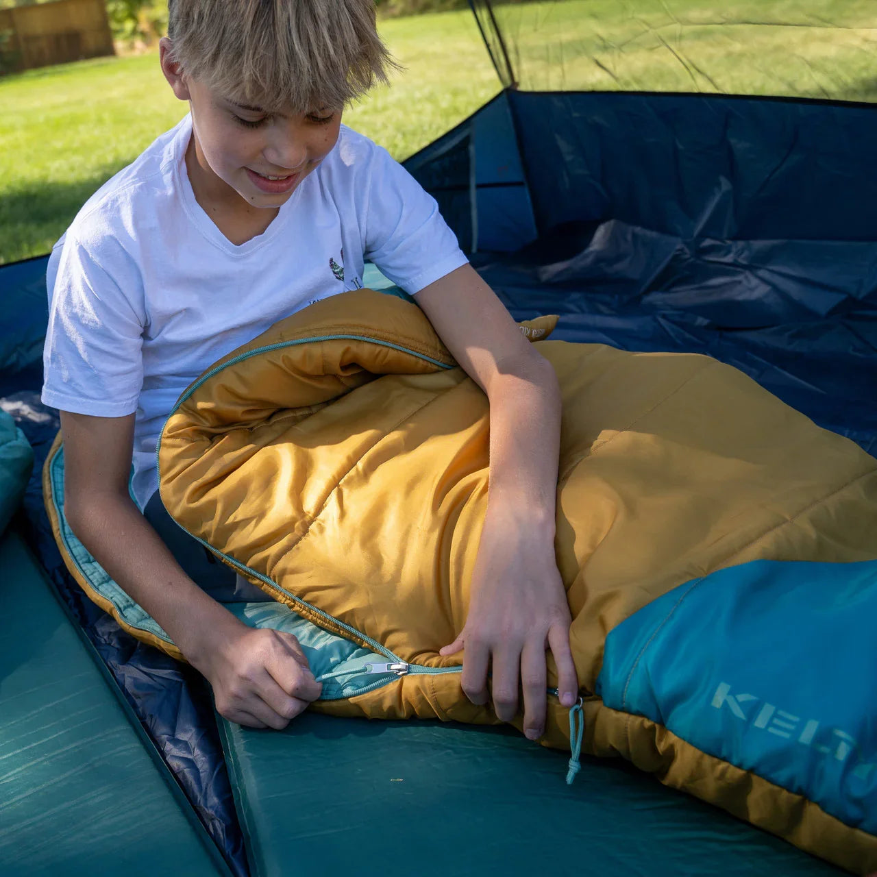 Boy in yellow and teal kids sleeping bag inside tent outdoors on a sunny day.