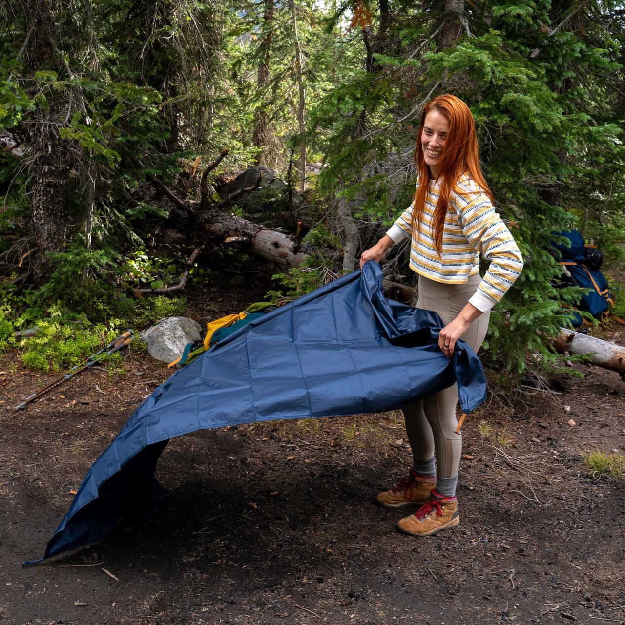 Woman setting up blue camping tarp in forest with hiking gear in background