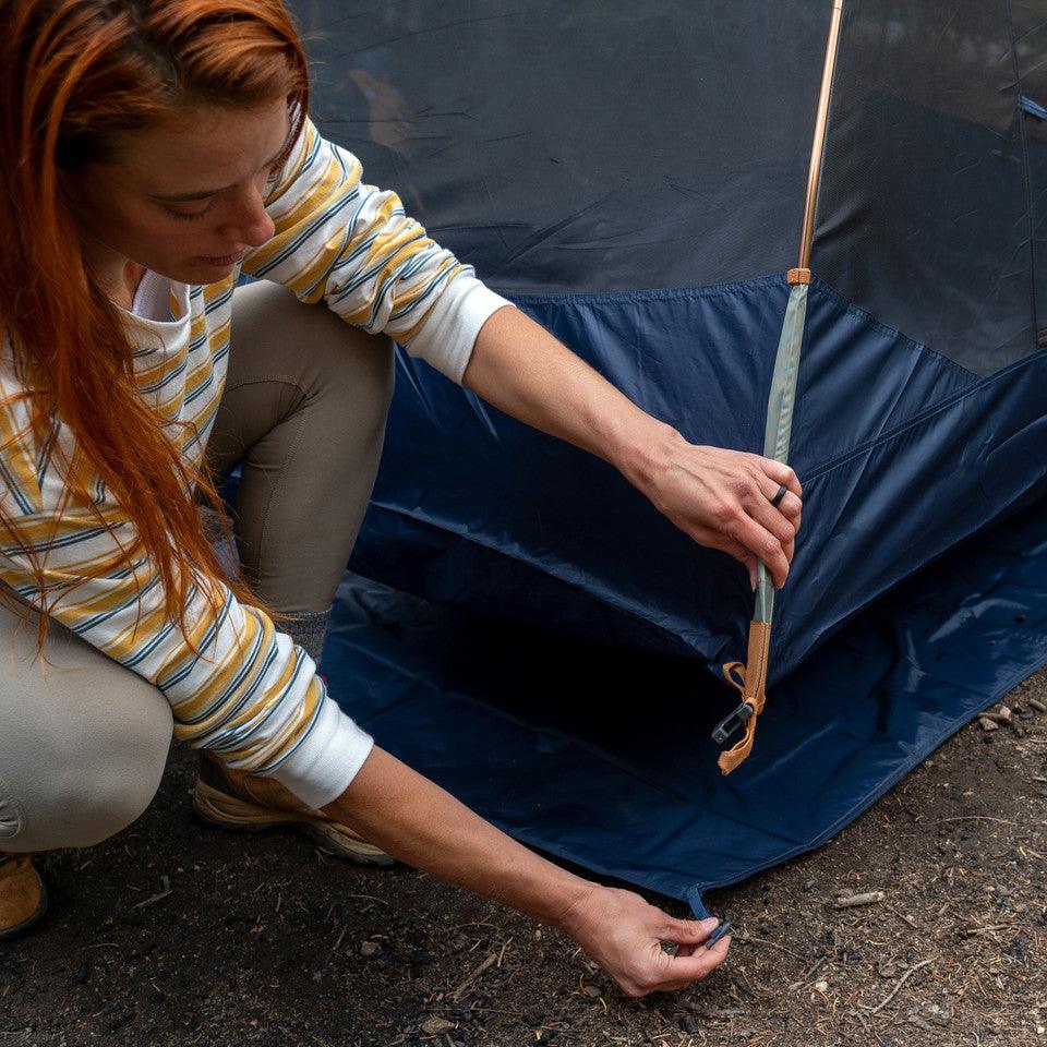 Woman setting up blue camping tent outdoors on dirt ground