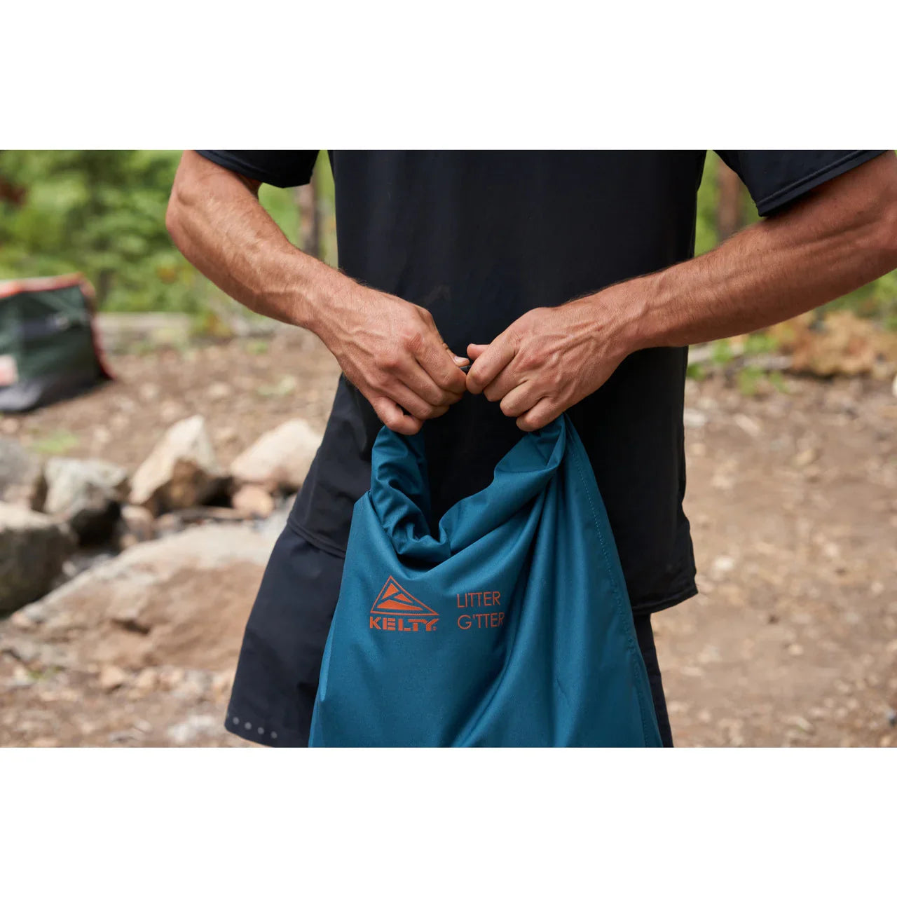 Person holding Kelty Litter Gitter deep teal bag outdoors in a rocky, wooded area