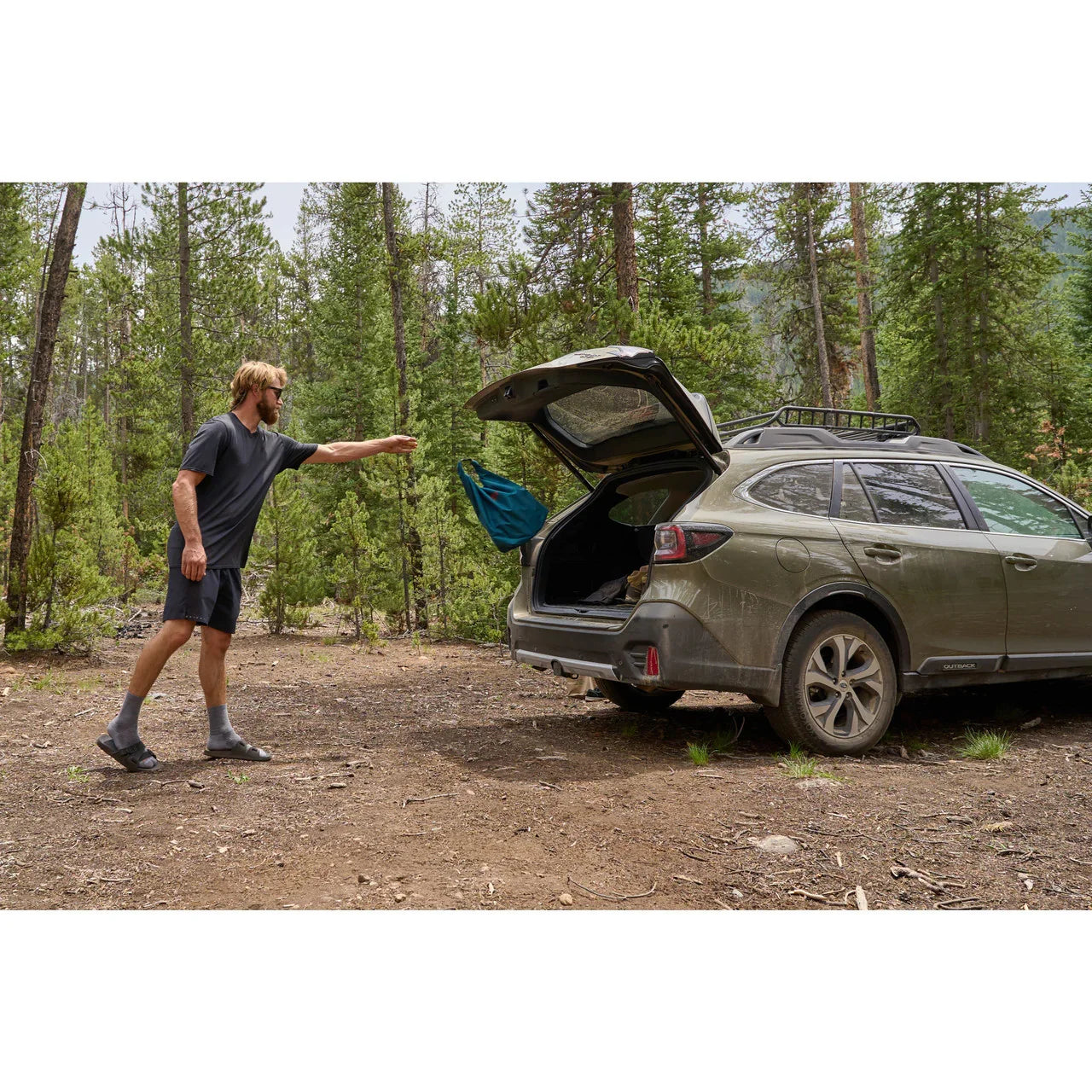 Man throws teal duffel bag into SUV trunk at forest campsite