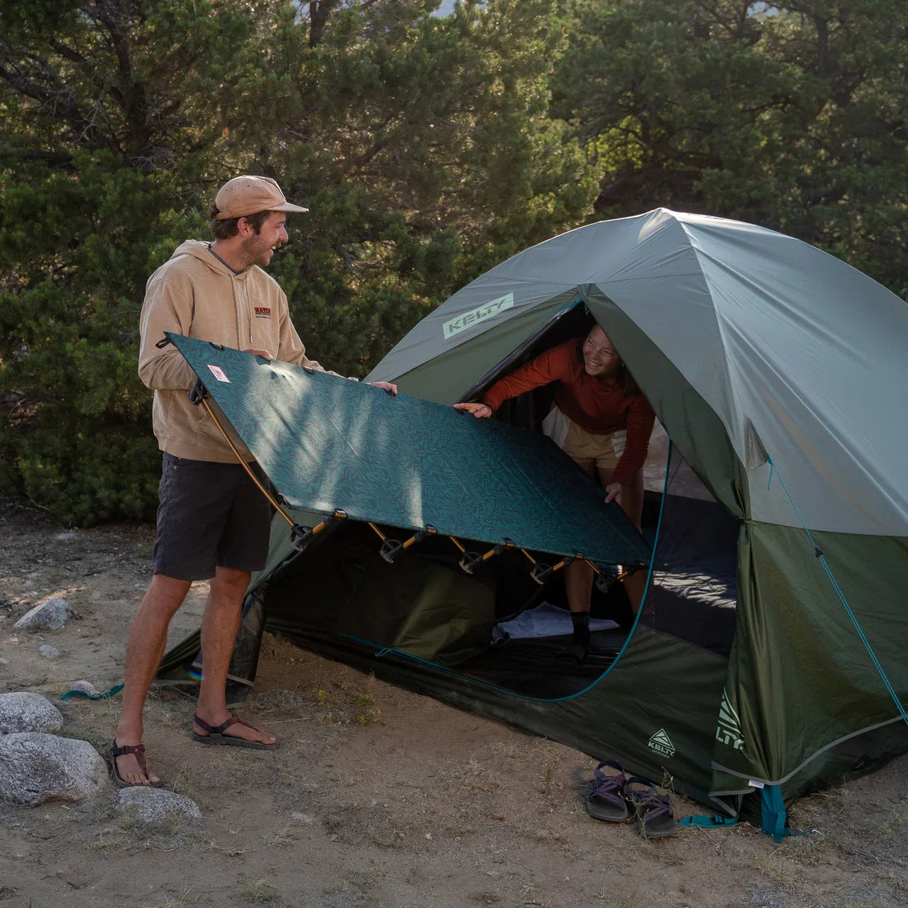 Two people setting up a camp cot outside a green Kelty tent in a forest campsite