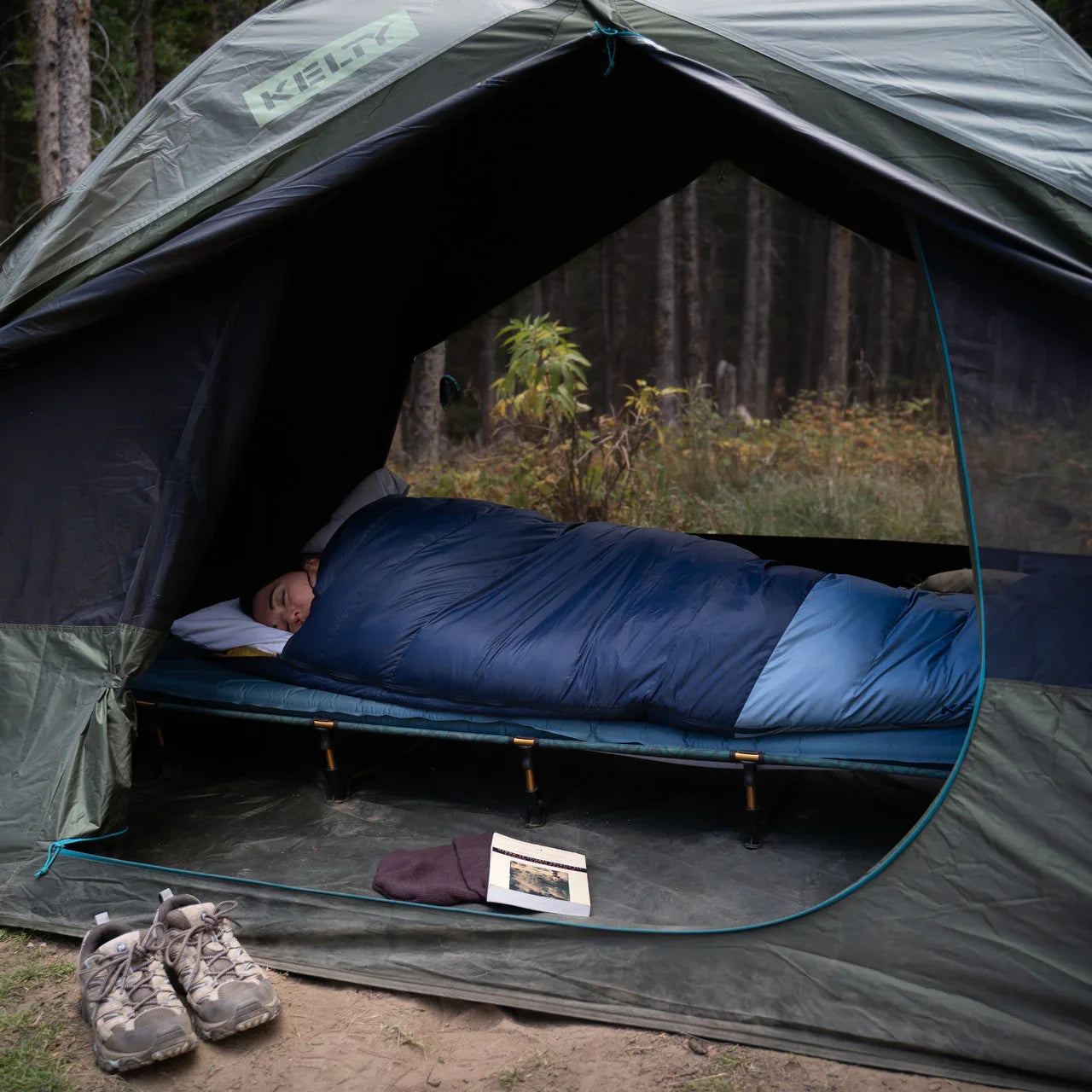Person sleeping in blue sleeping bag on camping cot inside tent with hiking shoes outside