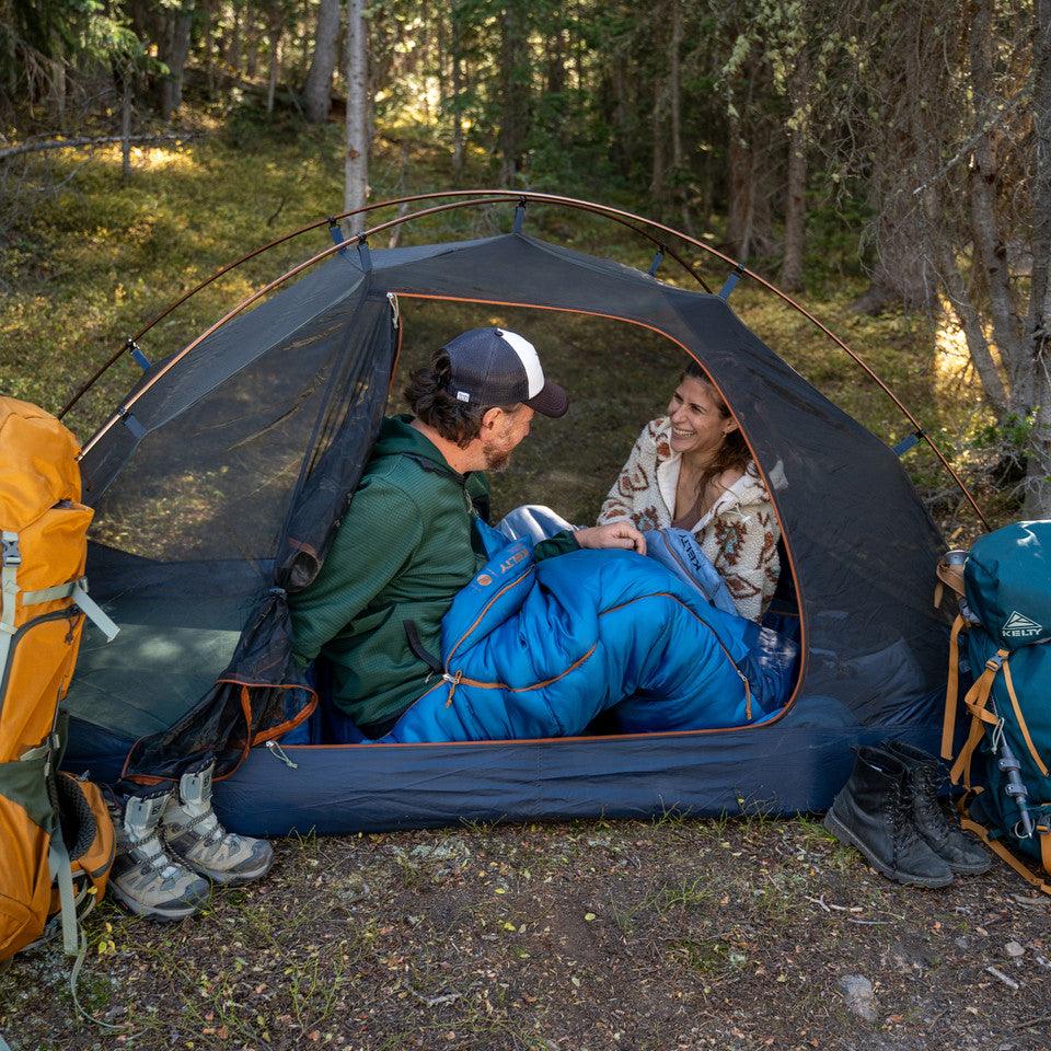 Two campers in a tent with sleeping bags in a forest, Kelty backpacks and hiking gear nearby