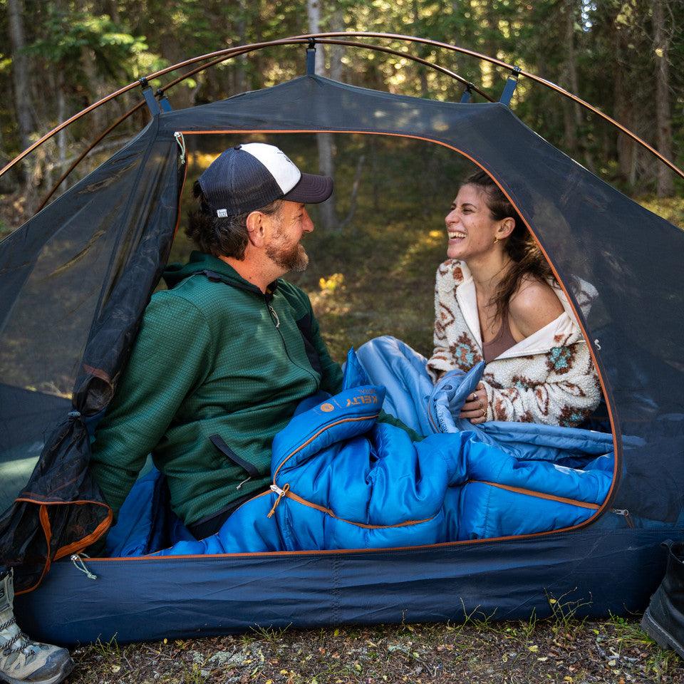 Couple smiling in a Kelty camping tent with blue sleeping bag in forest setting
