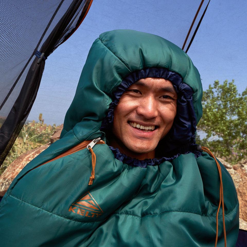 Smiling camper in green Kelty sleeping bag outdoors with trees and blue sky