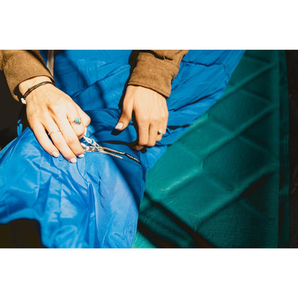 Hands zipping blue sleeping bag on green camping pad, outdoor gear close-up