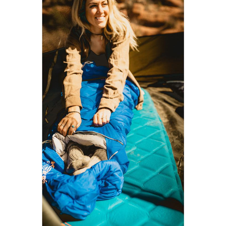 Woman camping in a blue sleeping bag on a teal sleeping pad outdoors
