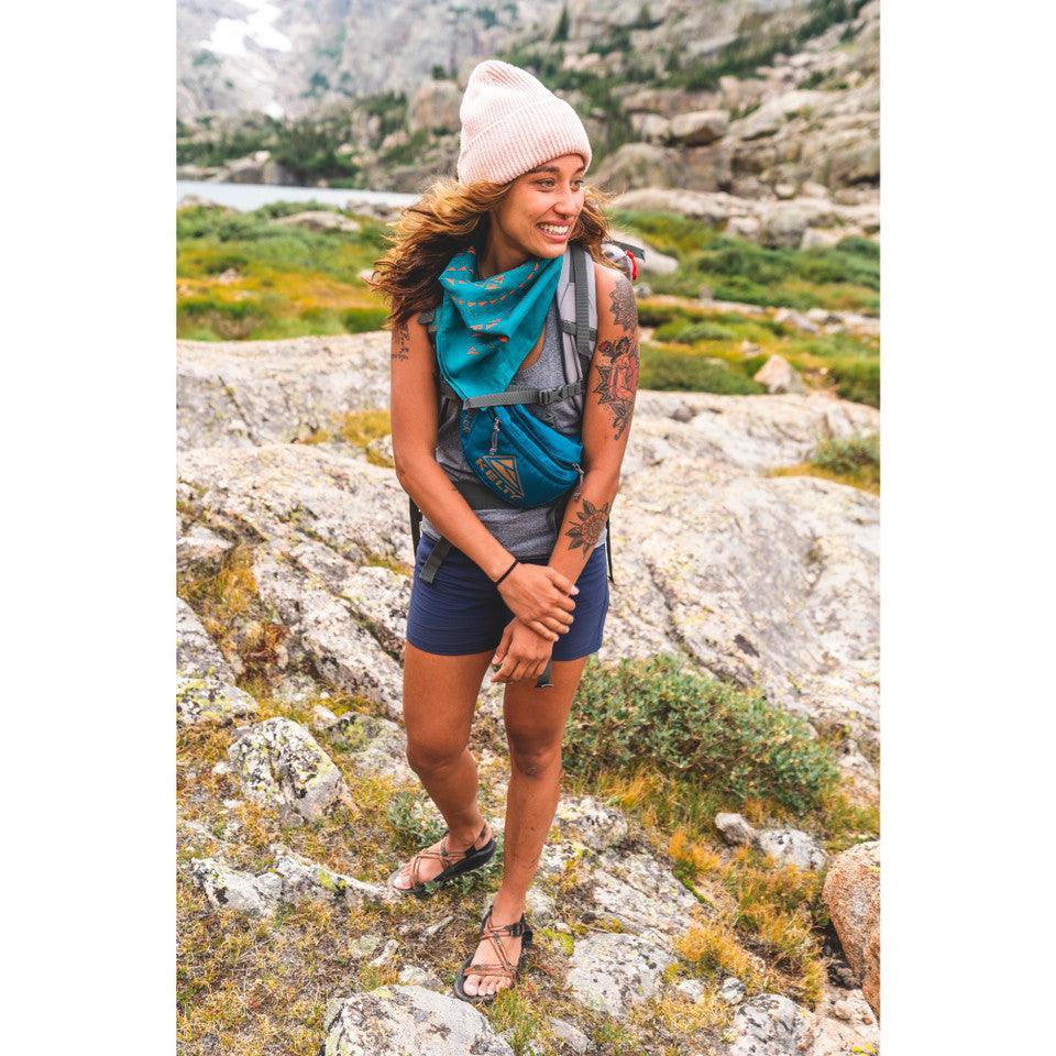 Woman hiking outdoors wearing Kelty Warbler fanny pack, blue bandana, shorts, and beanie