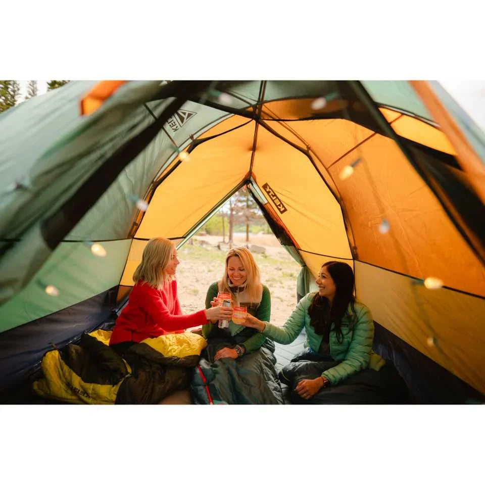 Three women in sleeping bags enjoy drinks inside a Kelty Wireless 2 camping tent outdoors