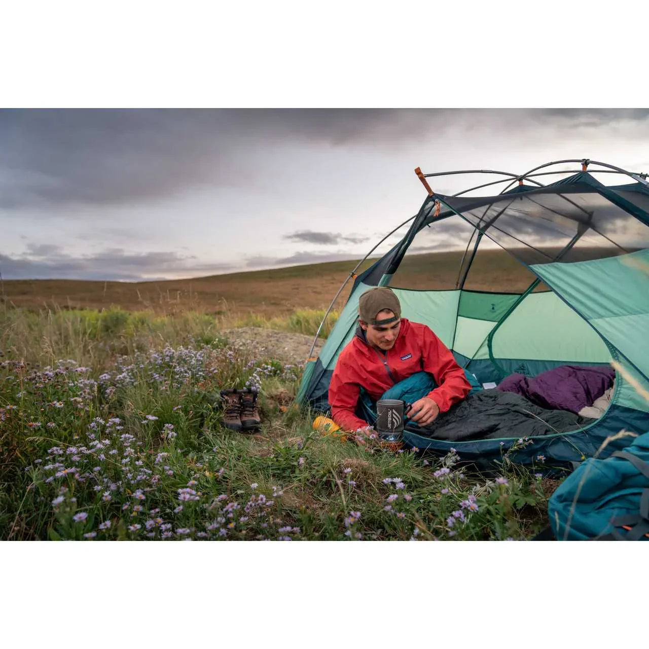 Camper in red jacket inside Kelty Wireless 2 Tent on grassy field with wildflowers at sunset