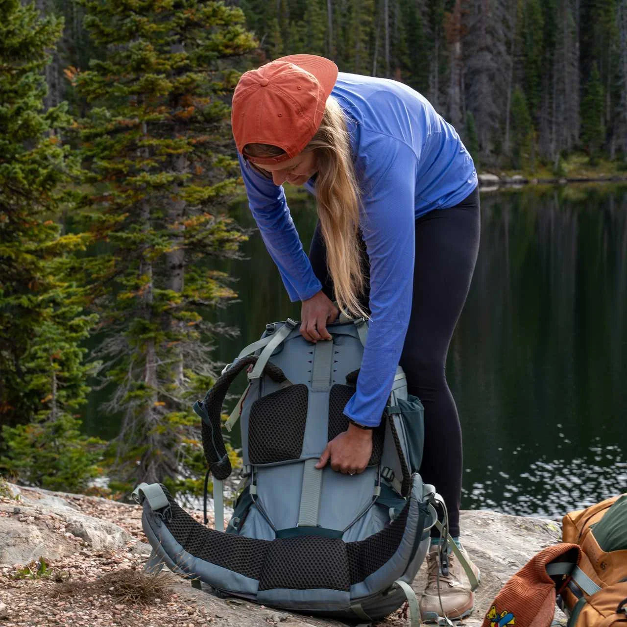 Woman outdoors adjusting Kelty Glendale 65L backpack by a forest lake