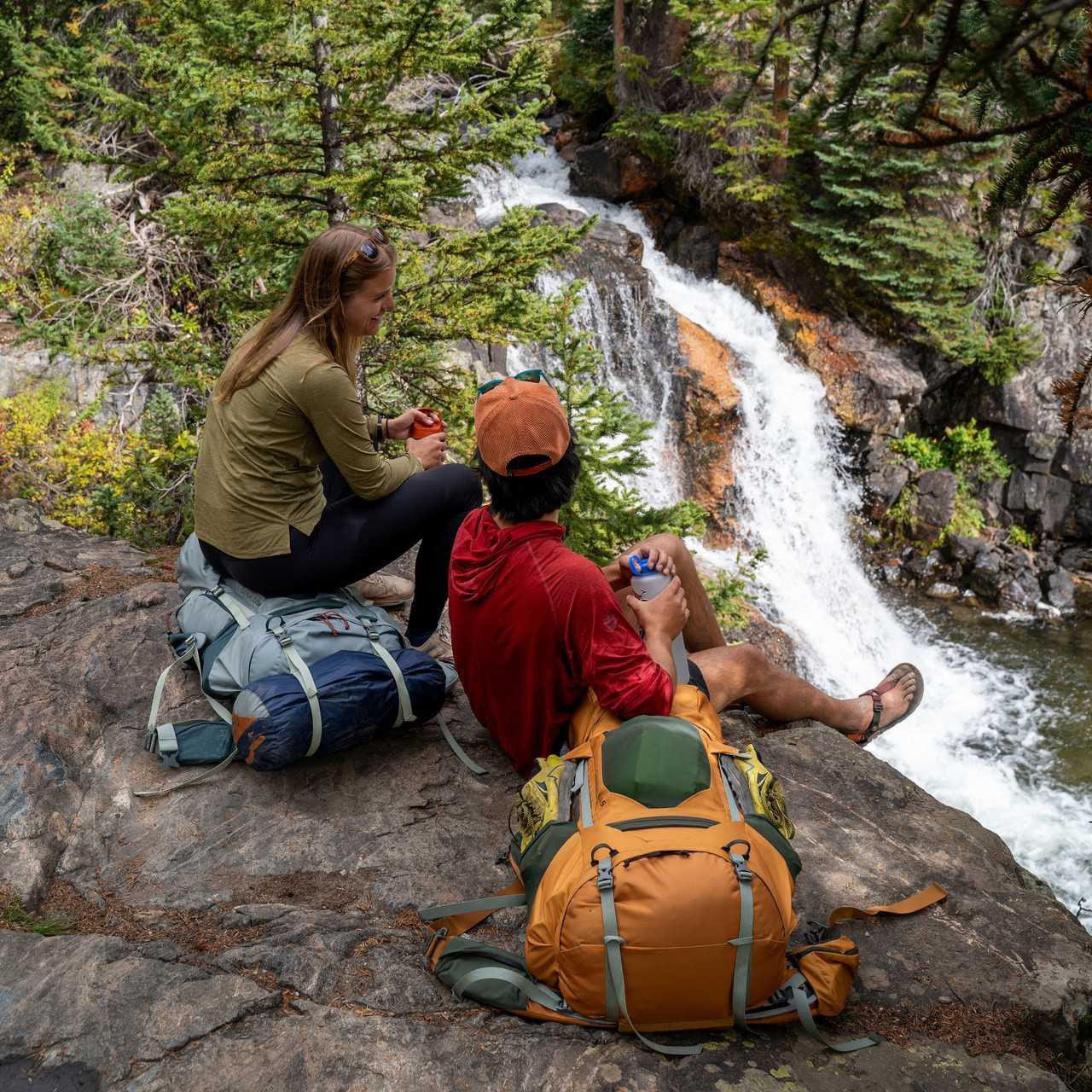 Two hikers with large backpacks rest on rocks by a forest waterfall, enjoying the view.