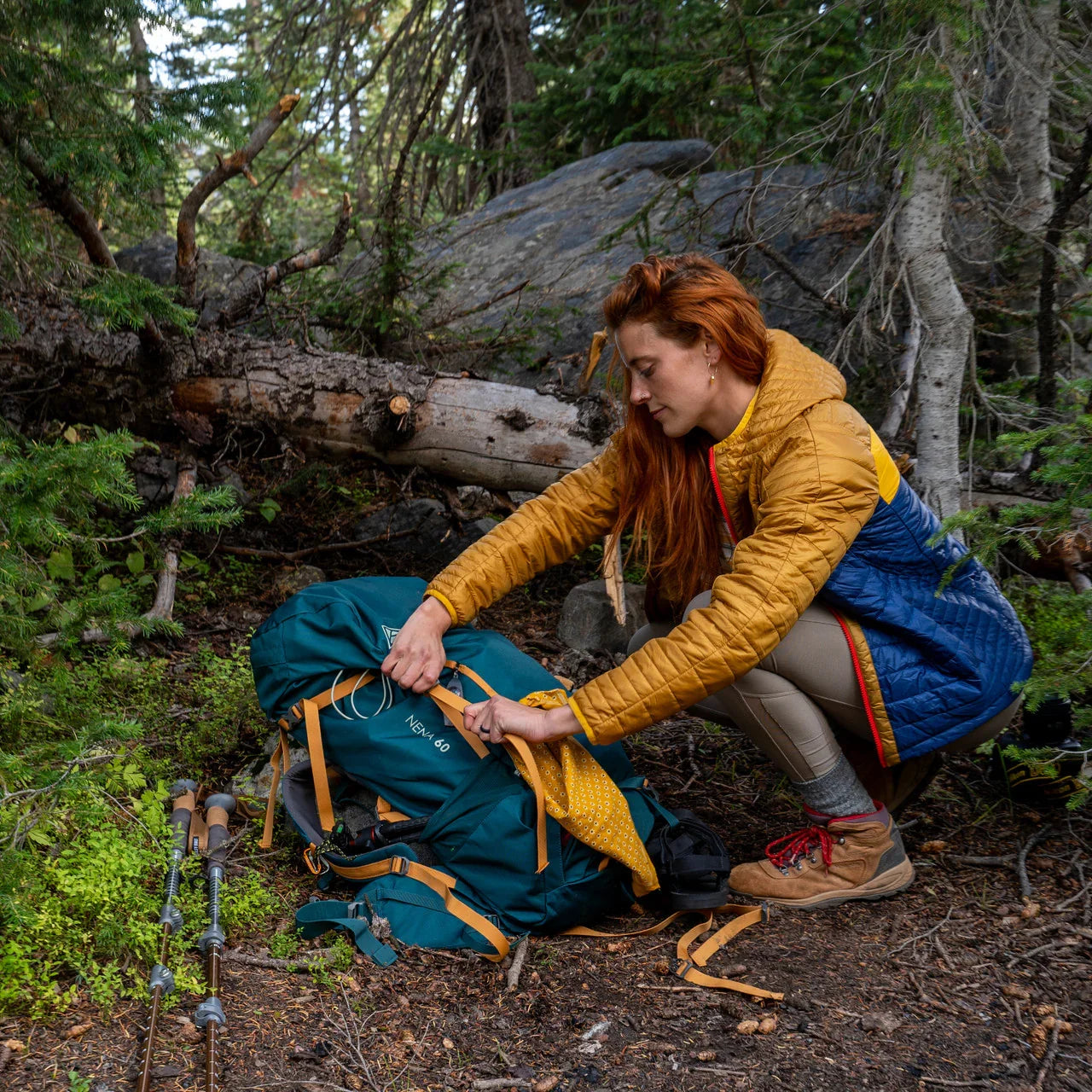 Woman hiking in forest adjusting teal backpack, wearing yellow and blue puffer jacket and boots